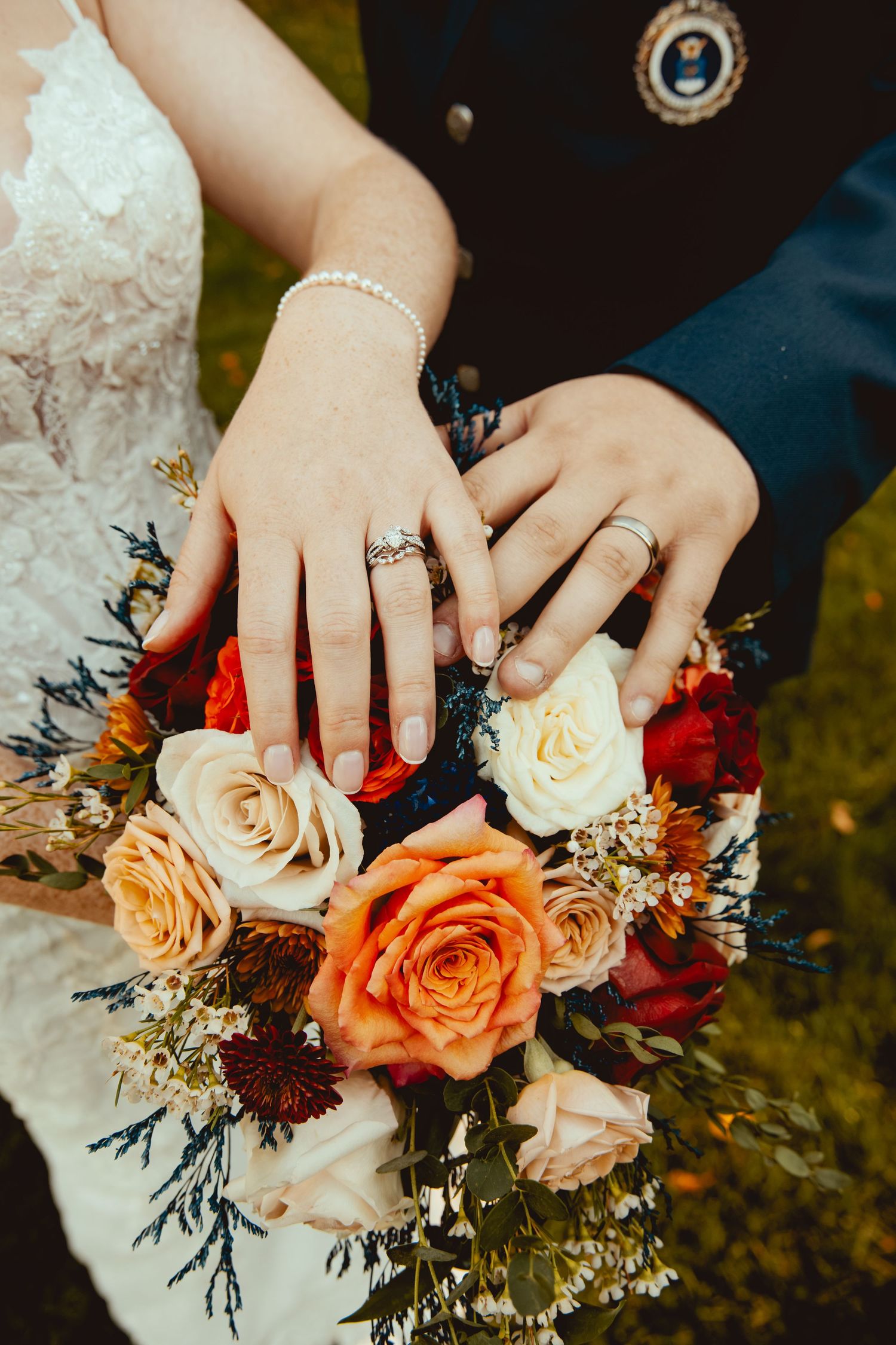 Newlyweds display wedding rings on a colorful bridal bouquet of orange, red, and cream roses.