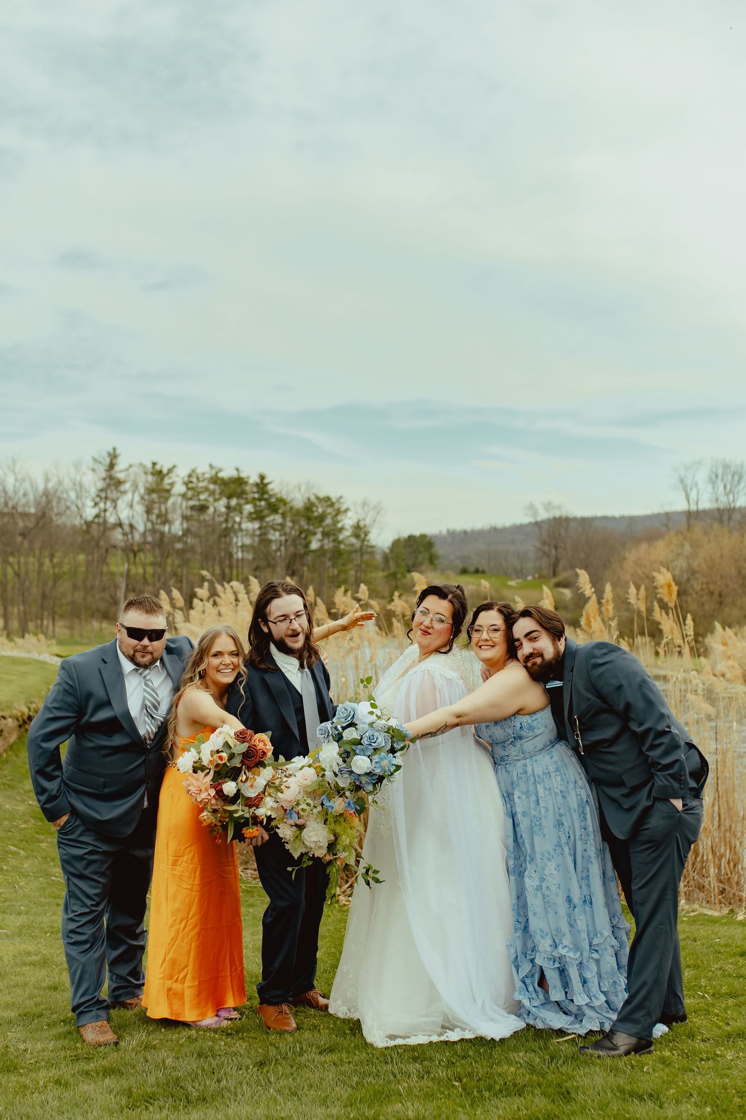 Bride in a white gown and wedding party in blue and orange attire laugh and embrace while holding bouquets in a grassy field.
