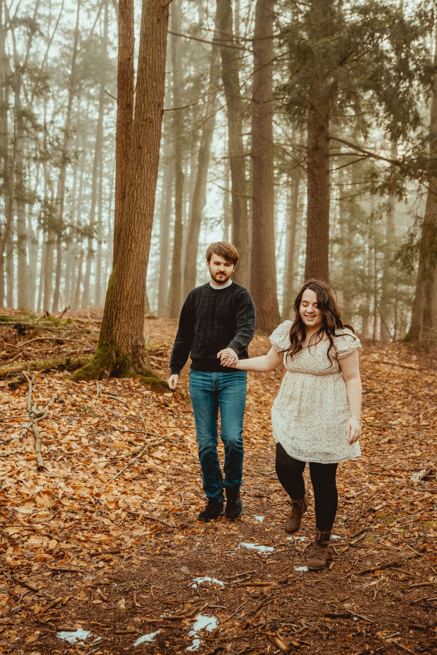 A couple strolls together through a foggy forest with fallen autumn leaves covering the ground.