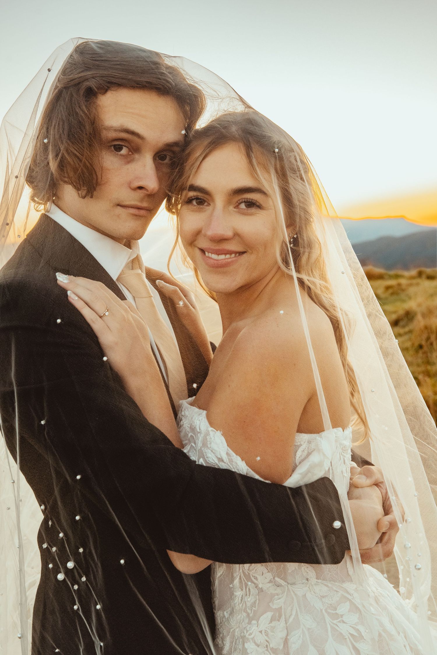 A bride and groom share a romantic moment under a pearl-adorned veil at sunset in a mountain landscape.