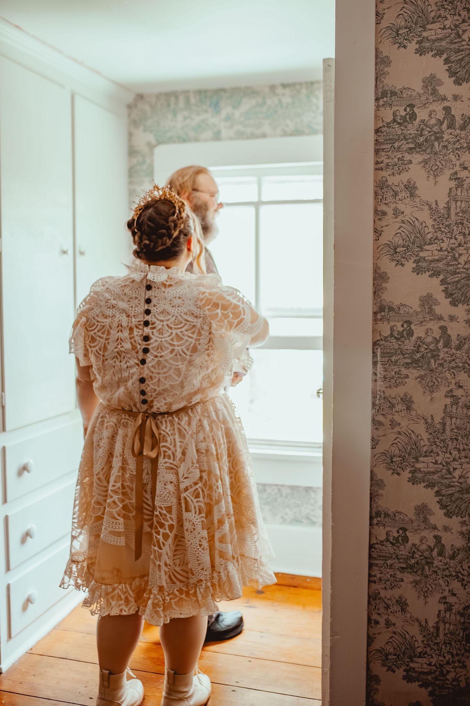 A person wearing a vintage-style lace dress with button details stands near a window in a room with floral wallpaper.