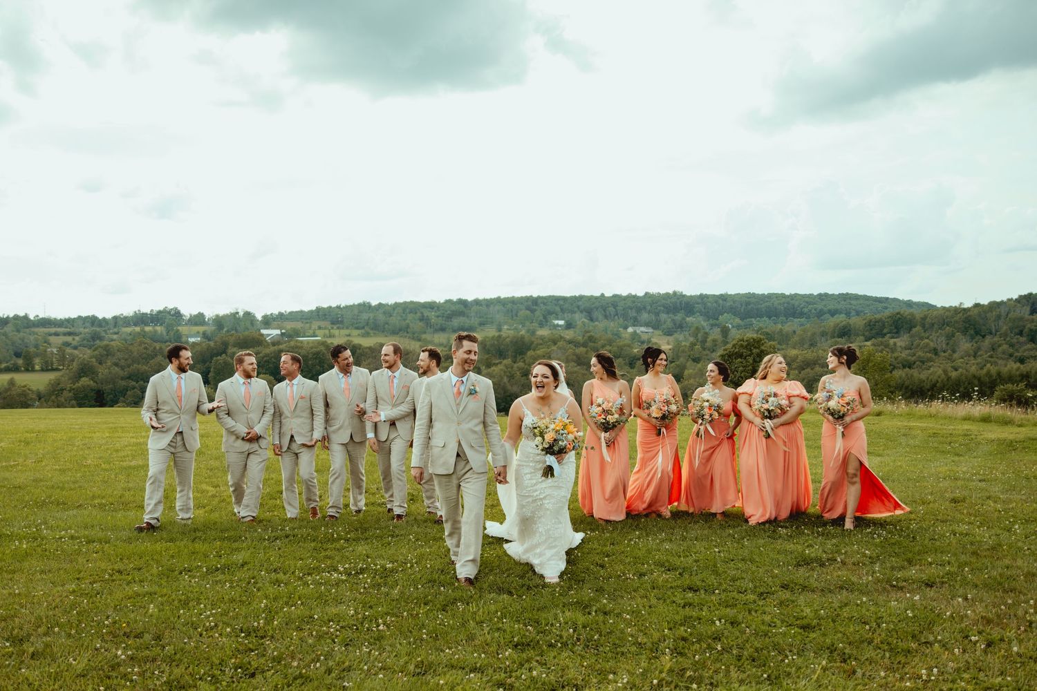 Wedding party walks in field, bridesmaids in coral gowns and groomsmen in tan suits.