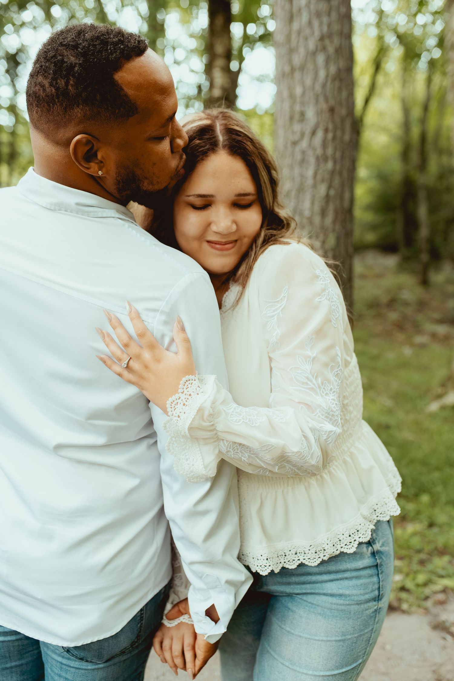 Couple sharing tender moment in forest, woman smiling as man kisses her cheek while embracing.