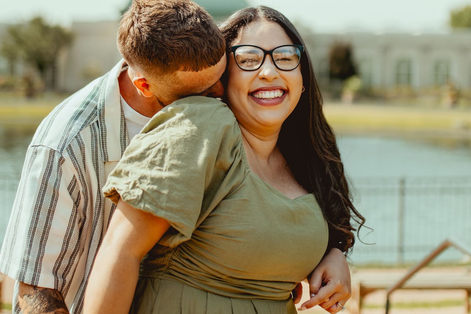 A smiling woman with glasses receives a kiss from a man outdoors near a lake, sharing a romantic moment.
