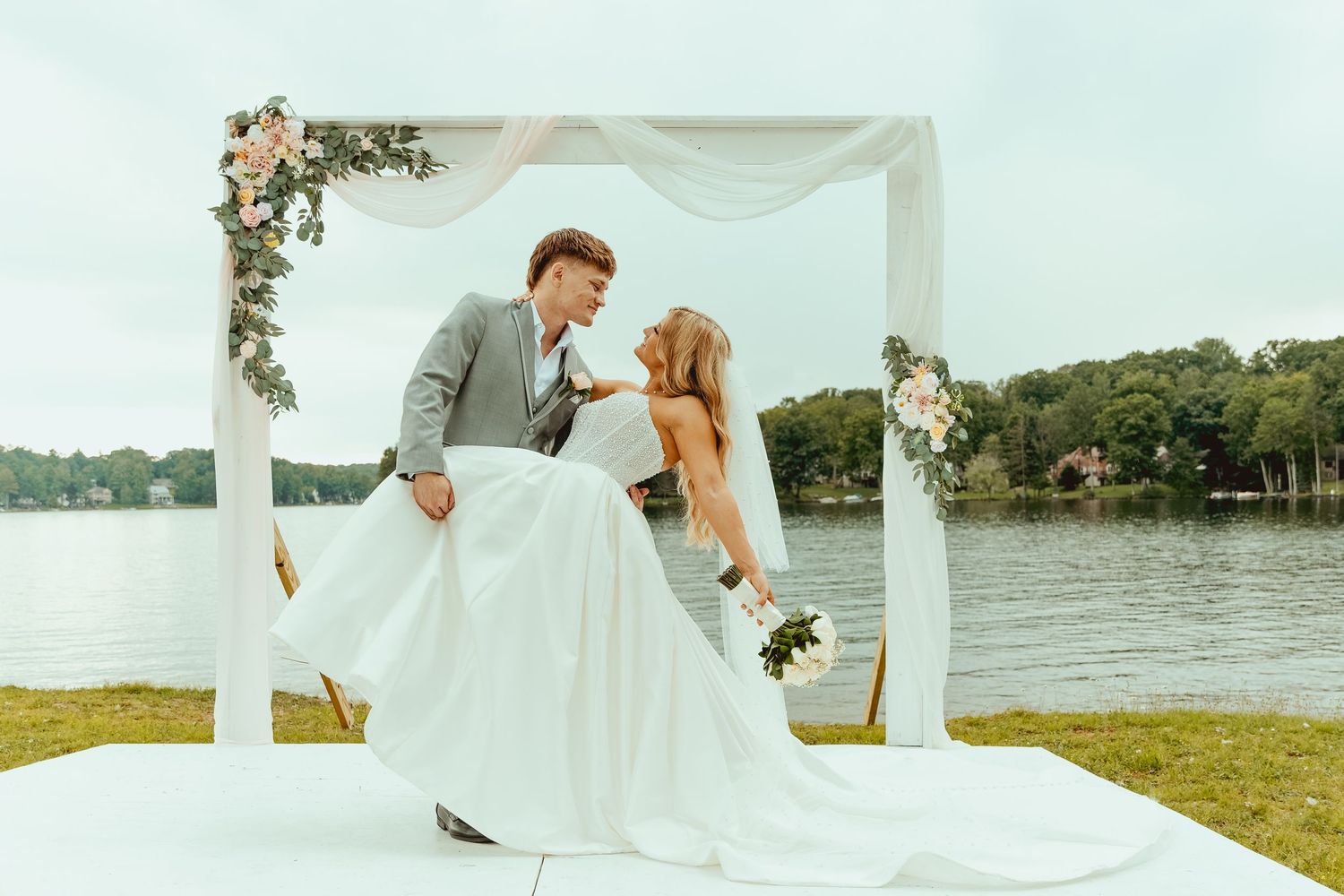 A couple shares a romantic kiss under a floral arch by a lakeside during their outdoor wedding ceremony.