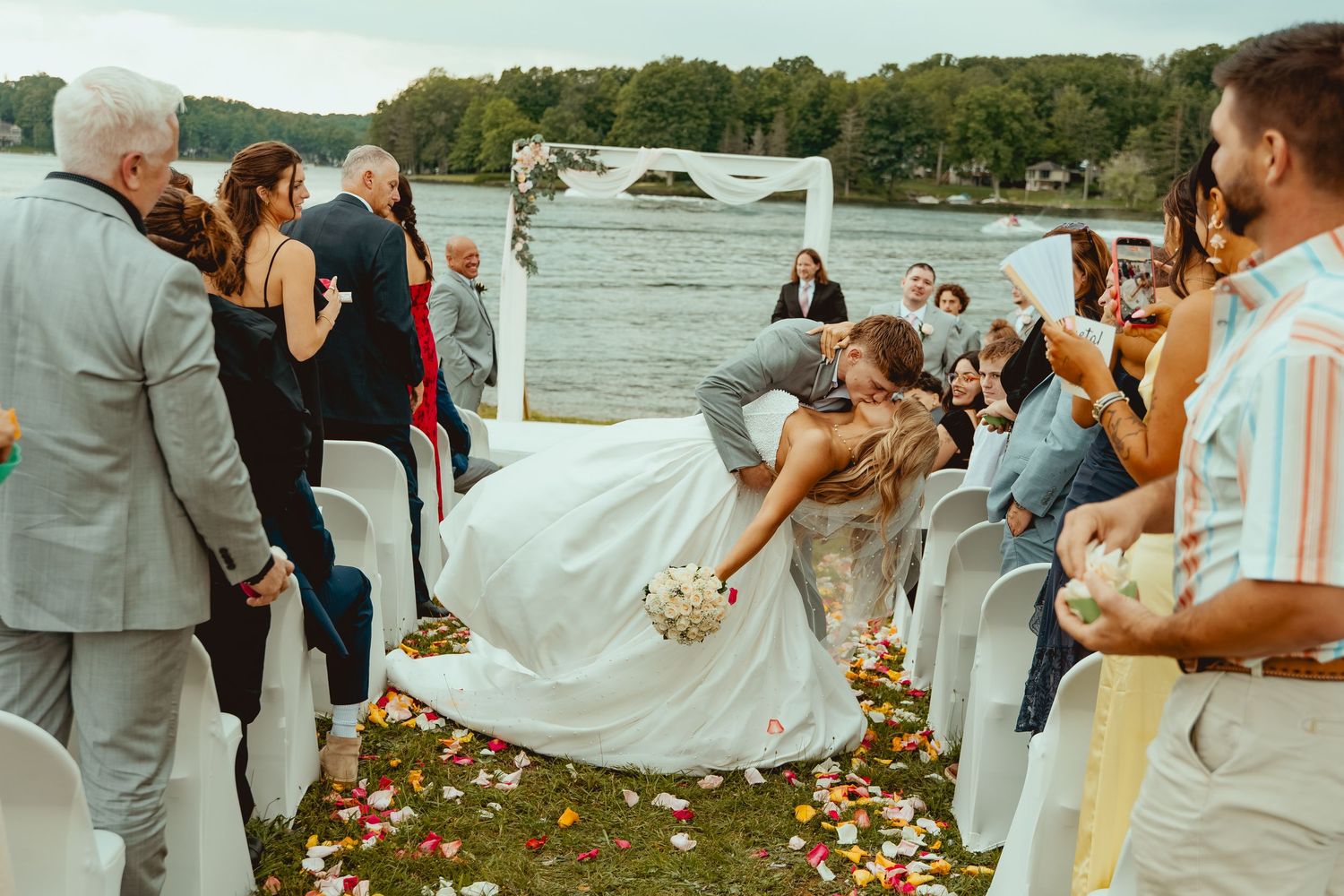 Bride and groom walking down aisle after wedding ceremony as guests applaud by waterfront with flower petals on ground.
