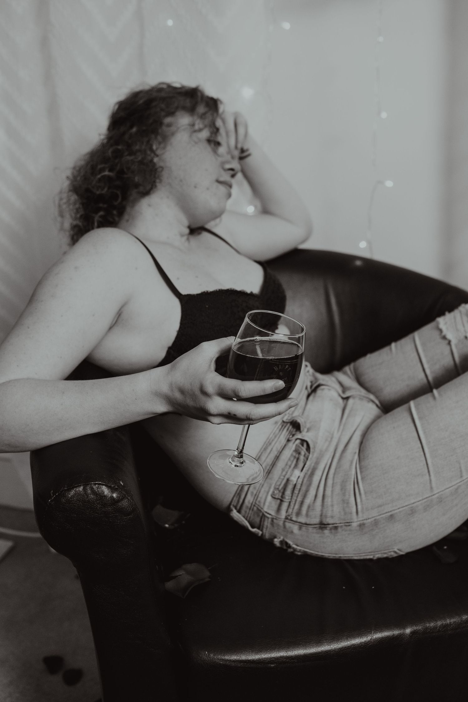 A black and white photograph shows someone reclining on a leather couch holding a wine glass while wearing casual attire.