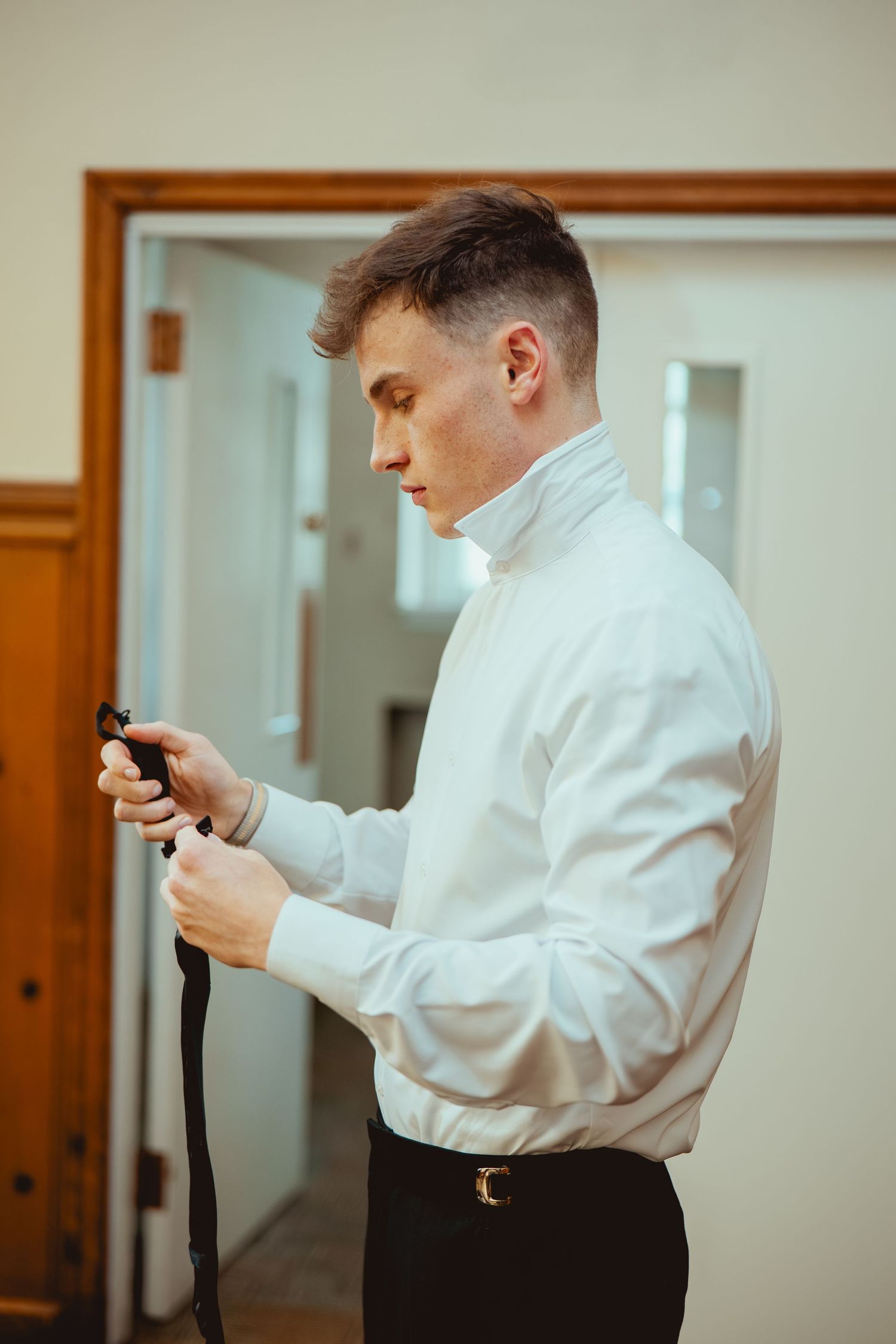A person in a white dress shirt adjusts their tie before a formal event in a warmly lit room.