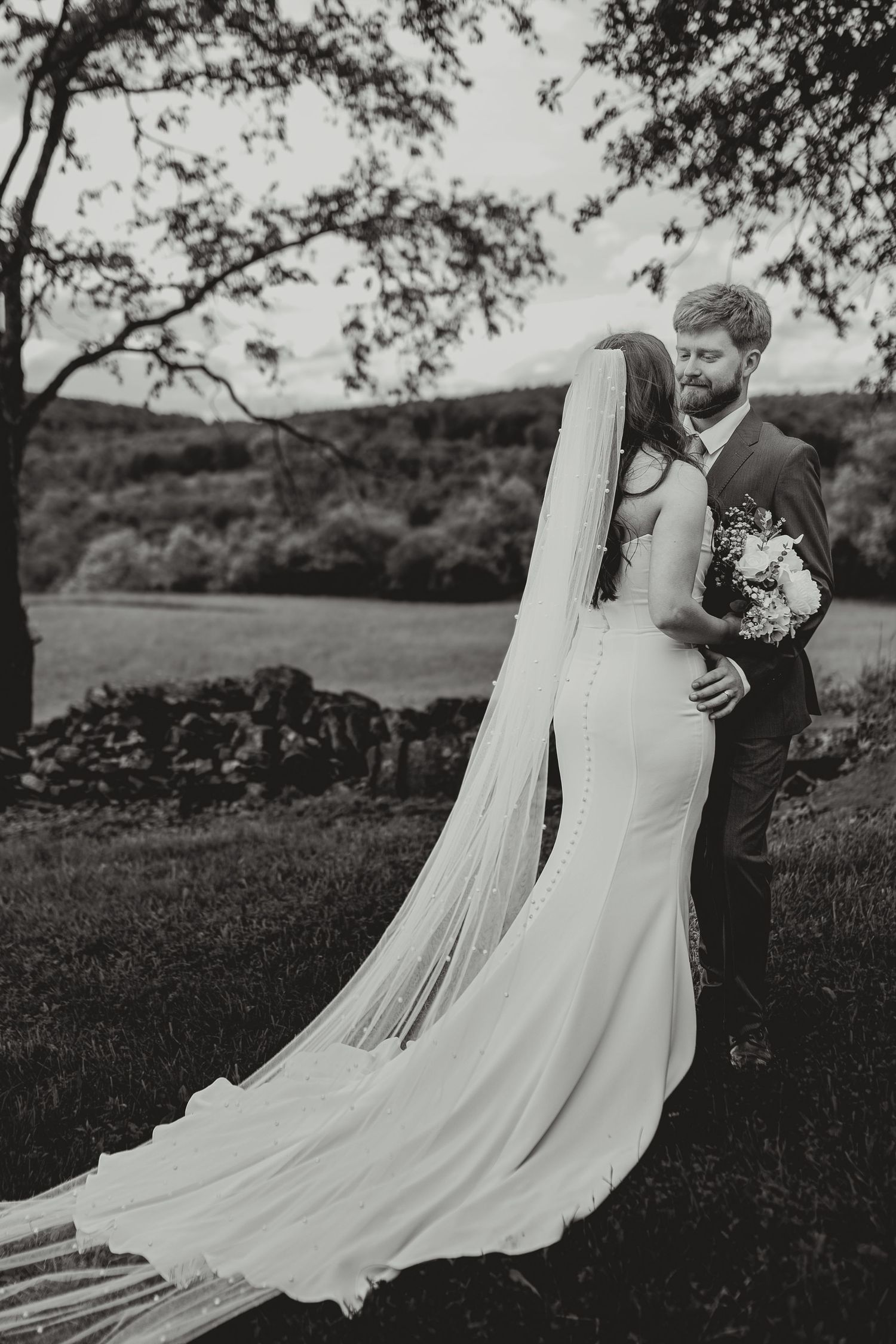Black and white wedding portrait with flowing veil against countryside landscape.