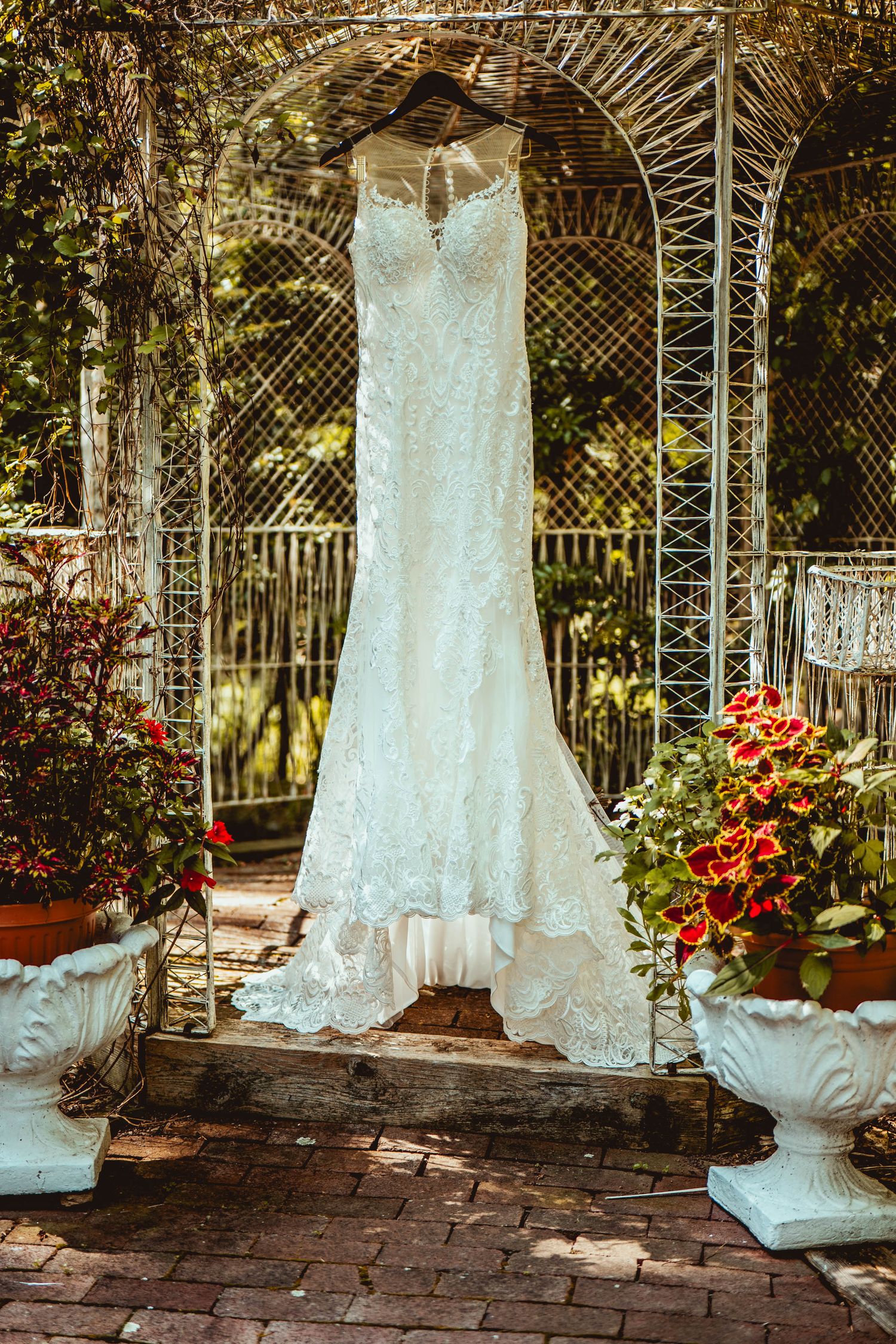 White lace wedding dress hanging in a garden gazebo with brick flooring and decorative floral arrangements.