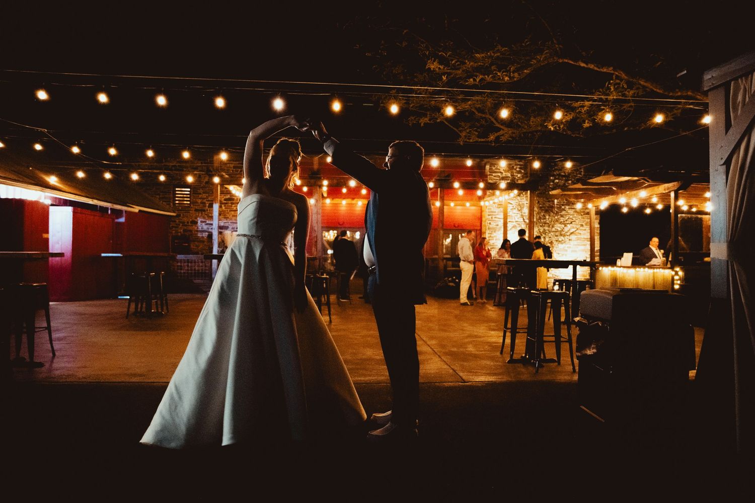 Bride and groom sharing romantic first dance under warm string lights at evening wedding reception.