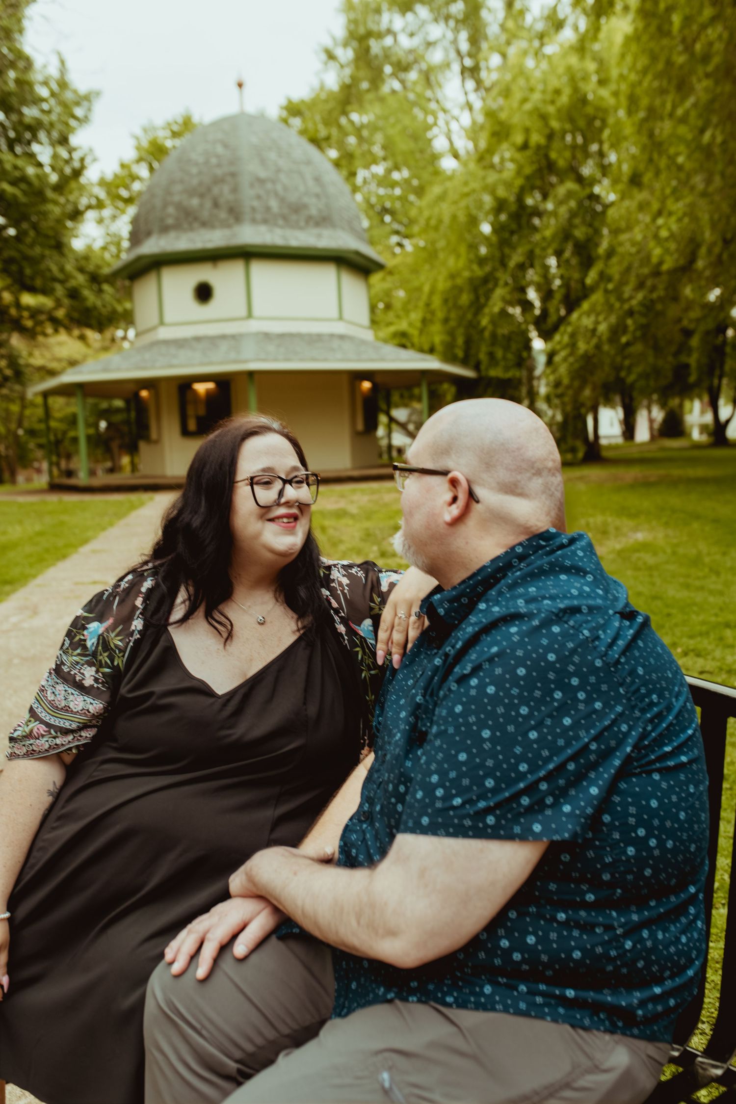 A couple sits on a bench in a park, smiling at each other with a gazebo and green trees in the background.