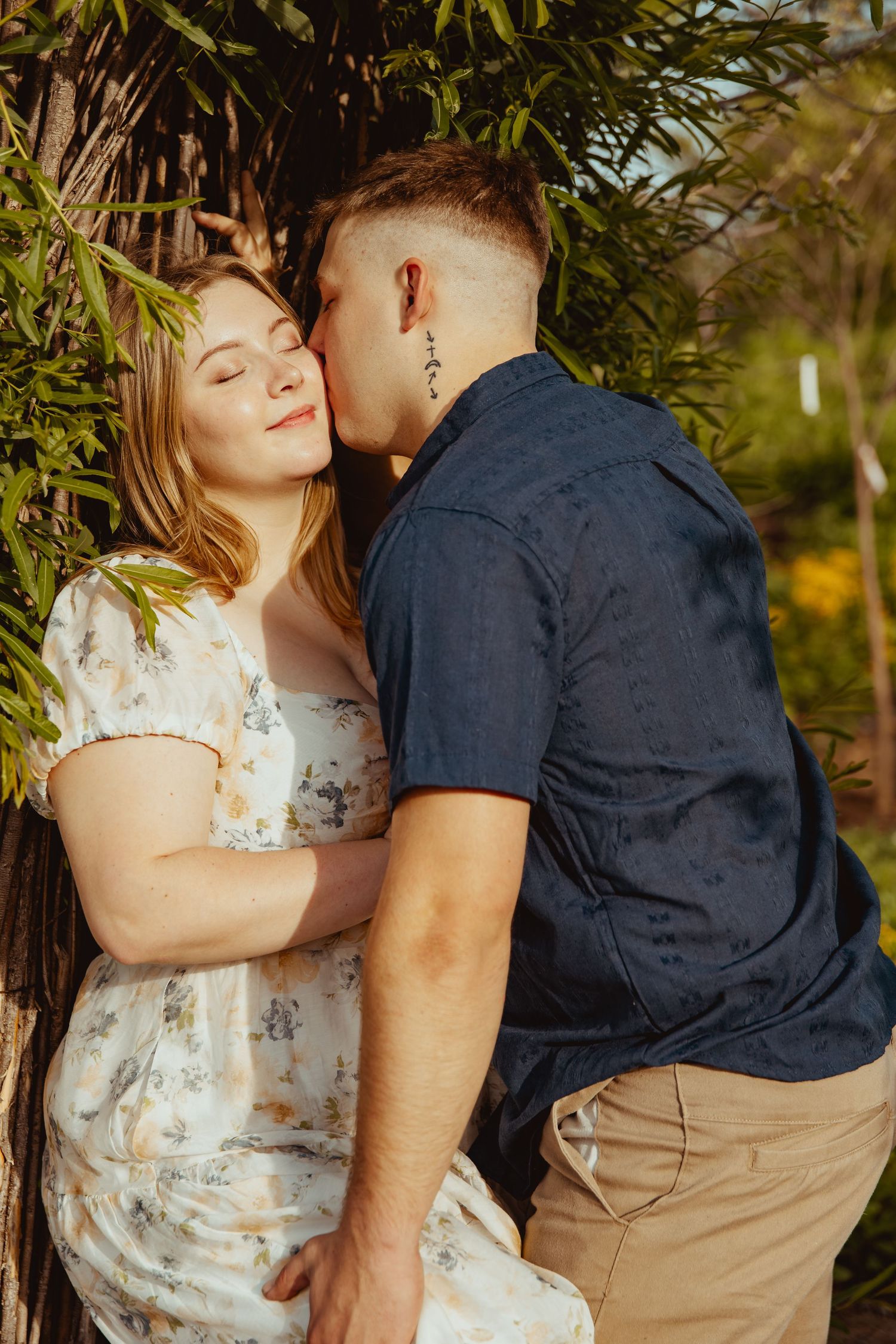 A couple shares a tender moment outdoors, man kissing woman's cheek near lush green trees in warm golden light.
