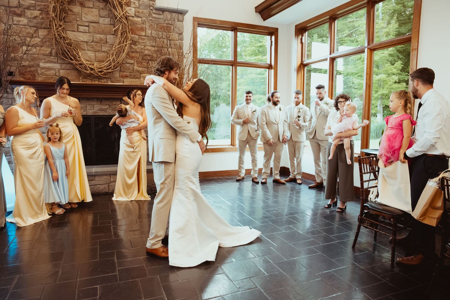 Bride and groom share first dance surrounded by wedding party in elegant rustic venue with stone fireplace.