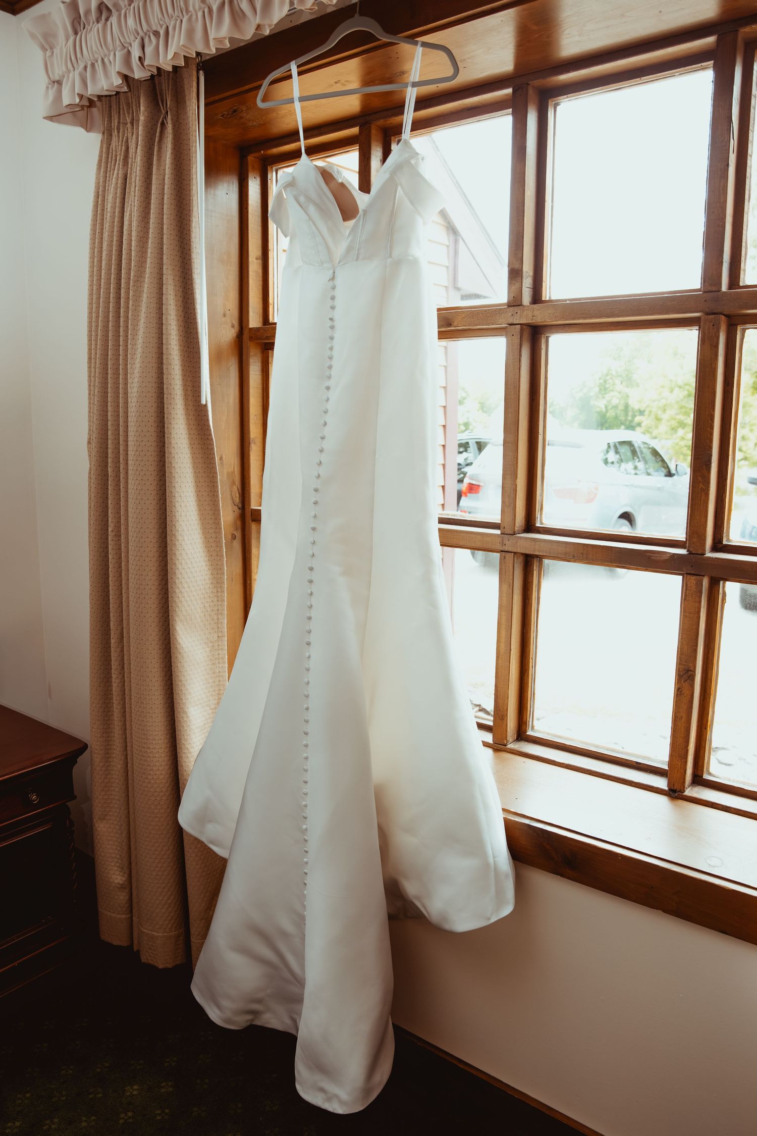 White satin wedding dress with button back detail hanging on wooden hanger in front of window.