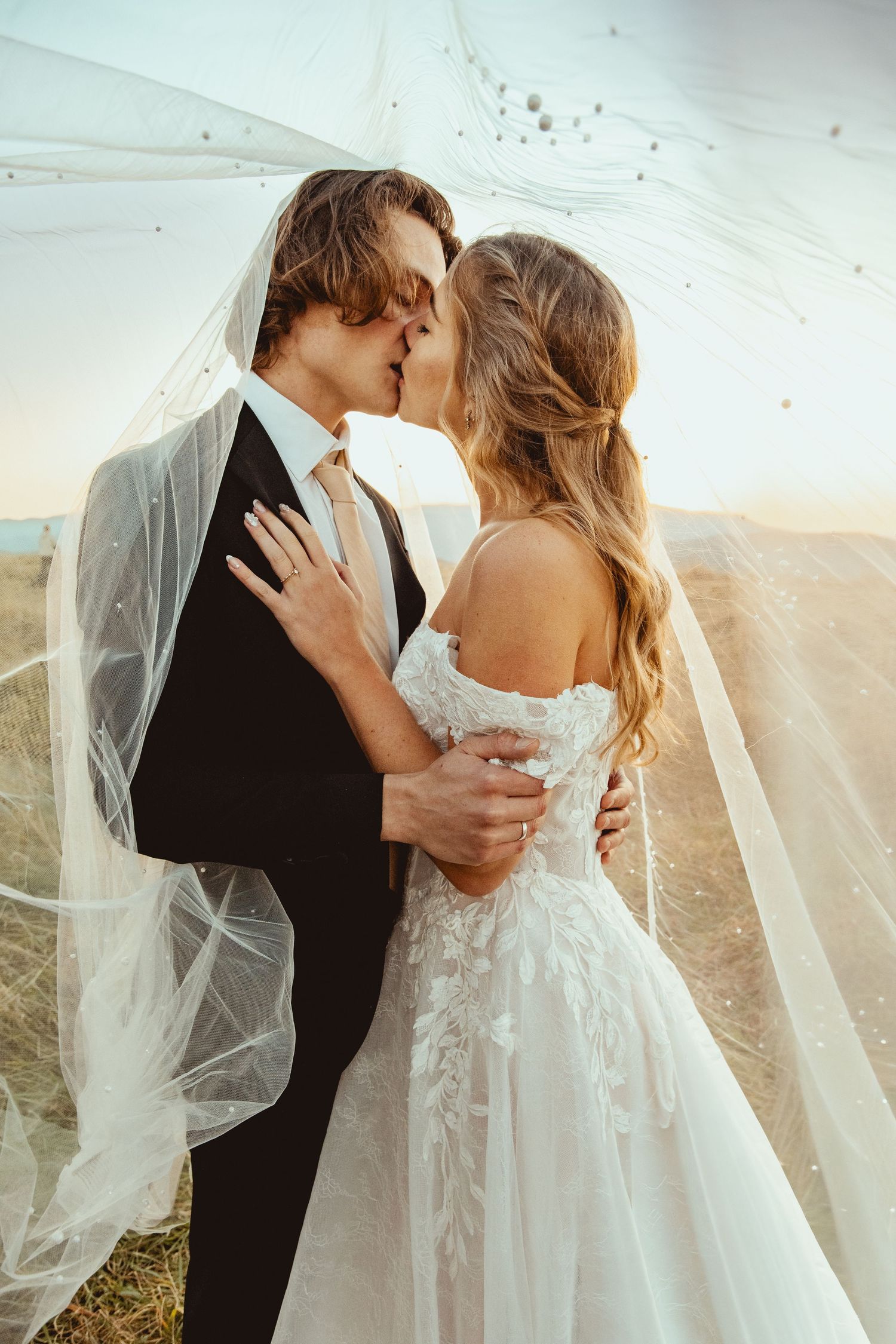 A romantic wedding couple embraces and kisses on a hilltop as a flowing veil catches the sunlight.