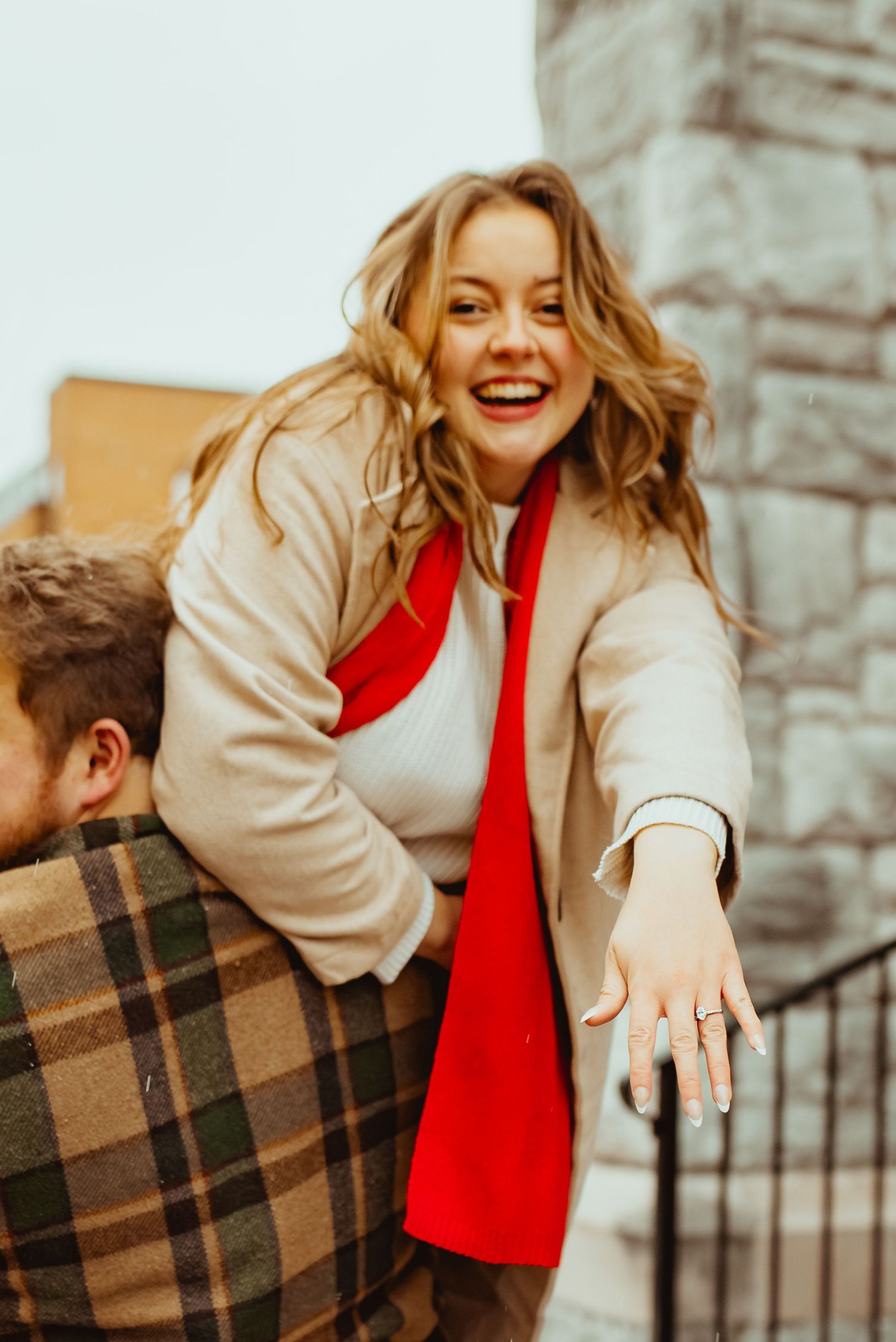 A couple shares a joyful moment together outdoors wearing a red dress and plaid coat in autumn lighting.