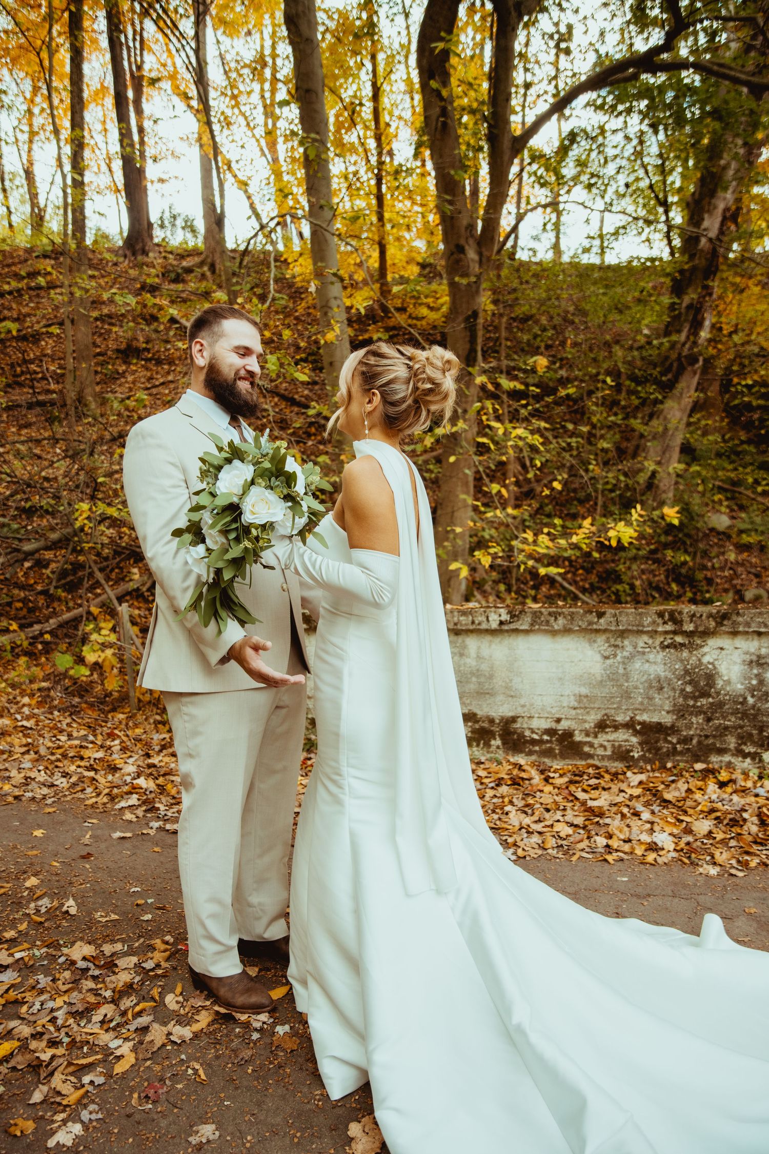 A couple in wedding attire shares an intimate moment in a scenic autumn forest setting with golden leaves.