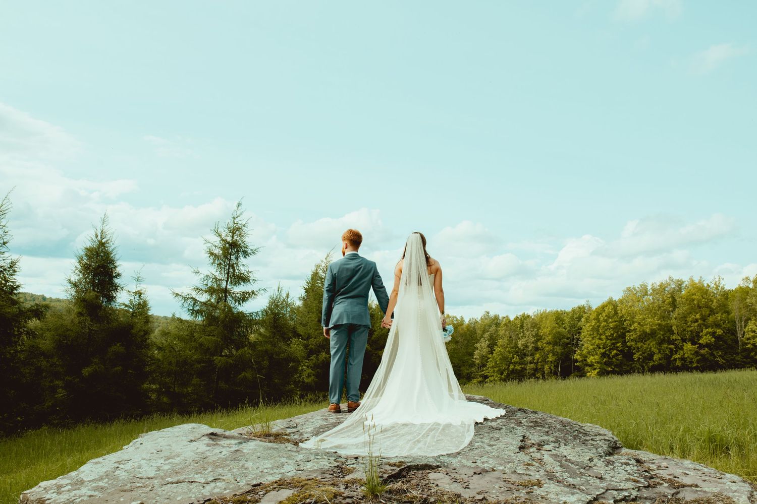 A couple in wedding attire walks through a scenic meadow with evergreen trees and blue sky in the background.