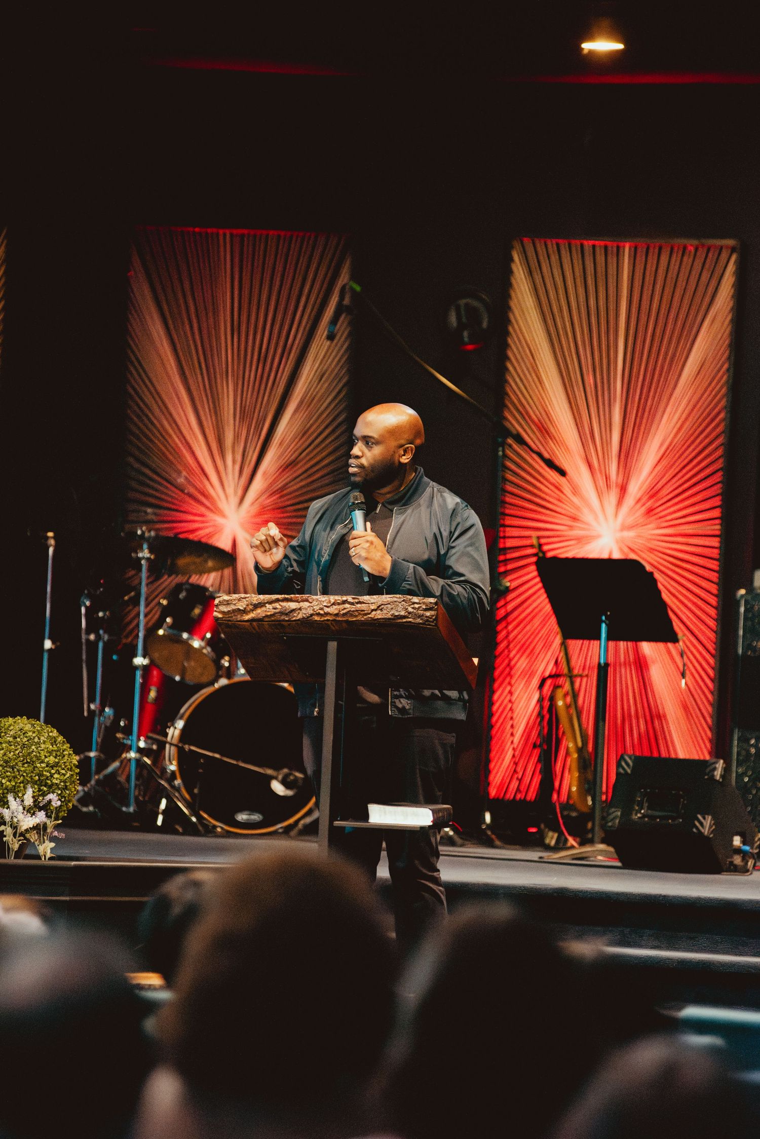 Speaker stands at podium on stage with dramatic red sunburst backdrop panels and musical instruments.