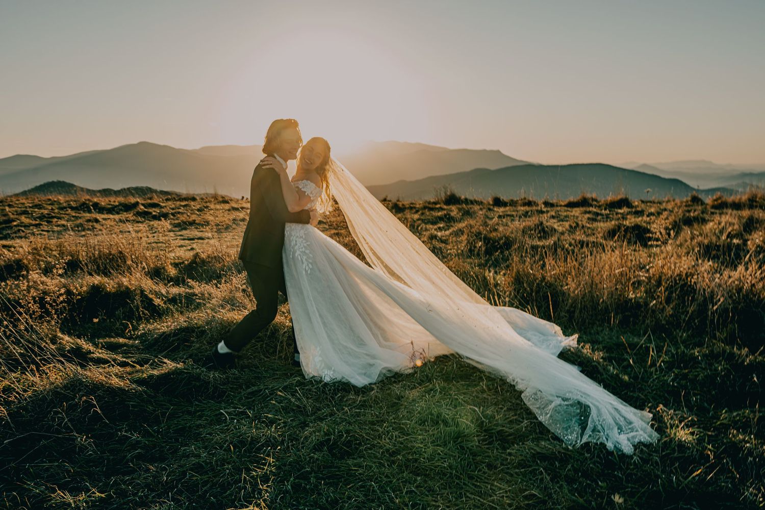 A couple in wedding attire embrace on a hilltop during golden hour as the bride's veil flows dramatically in the wind.