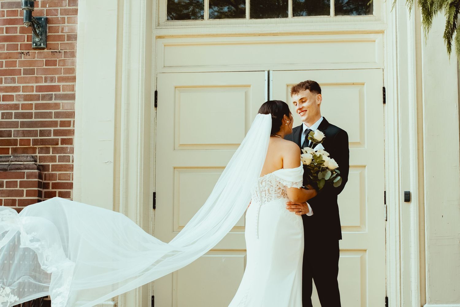 A bride and groom share a romantic moment in front of a white door and brick wall during their wedding photoshoot.