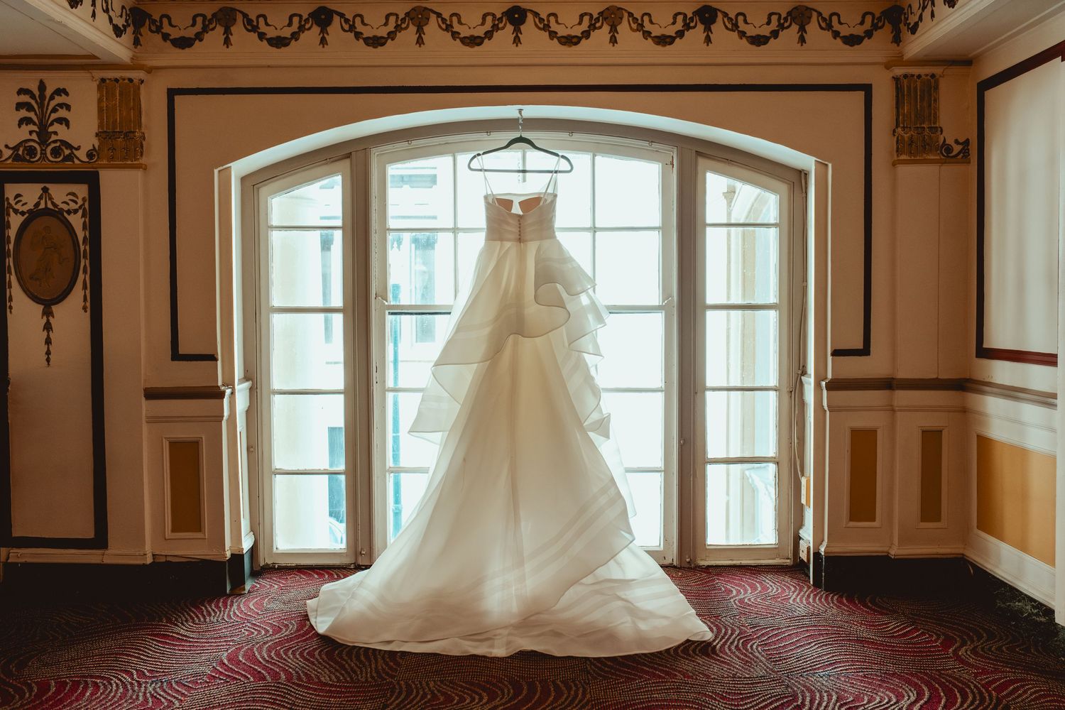 Elegant white wedding dress displayed in a grand room with ornate architectural details and large windows.