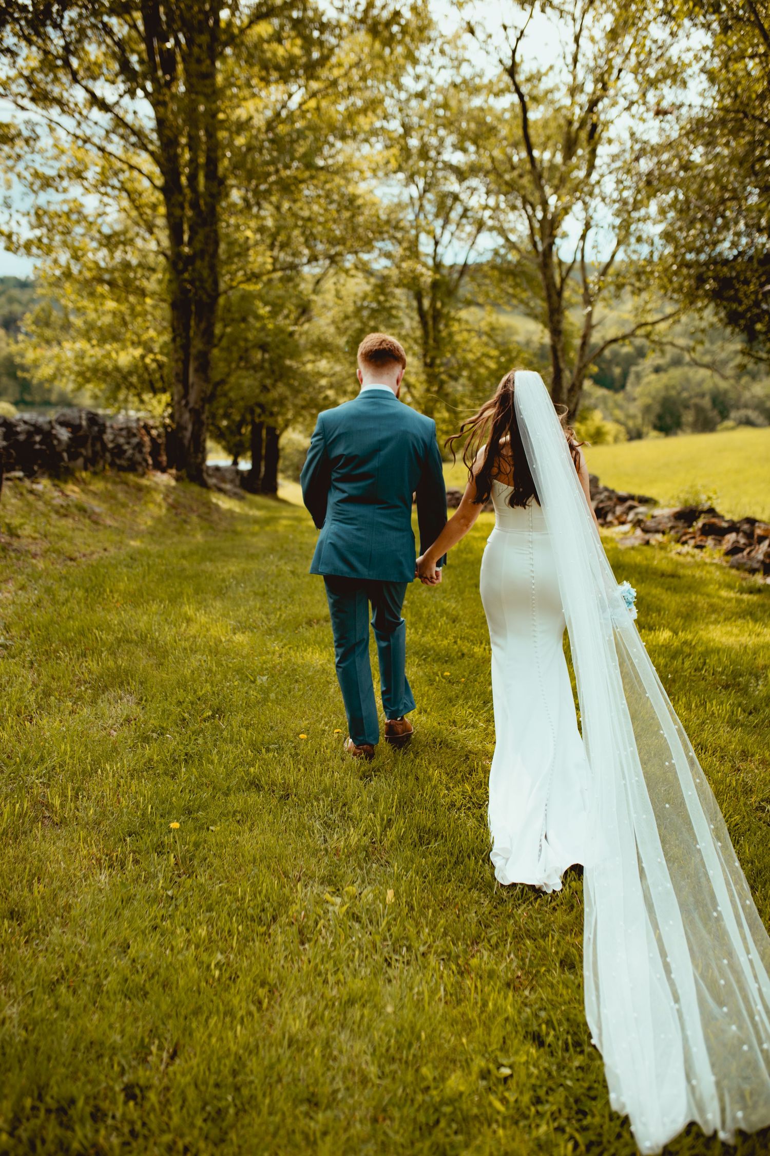 A couple walks hand in hand through a sunlit meadow with stone walls and trees in the background.