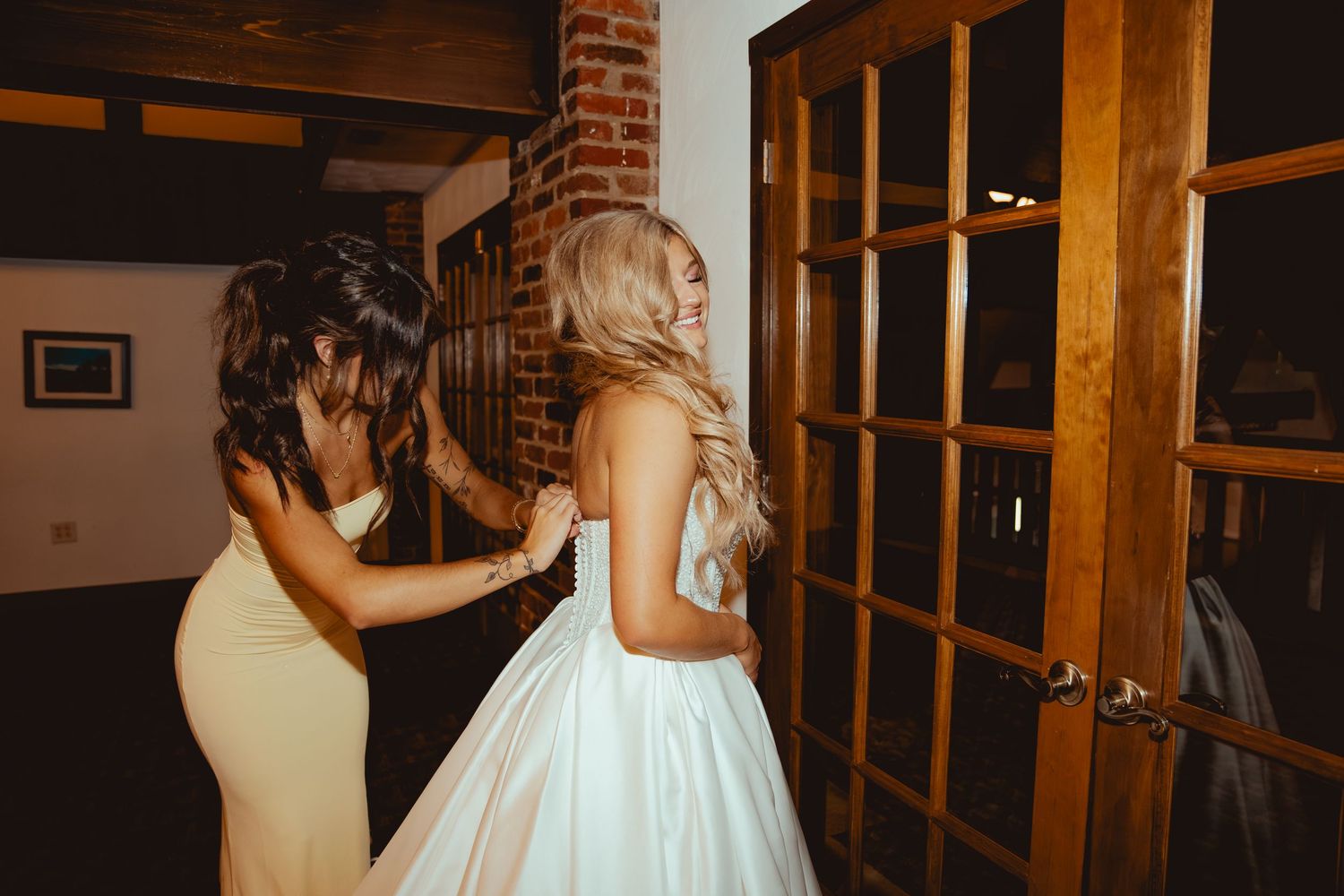Bridesmaid in yellow dress helping bride in white ball gown with her dress near wooden French doors.