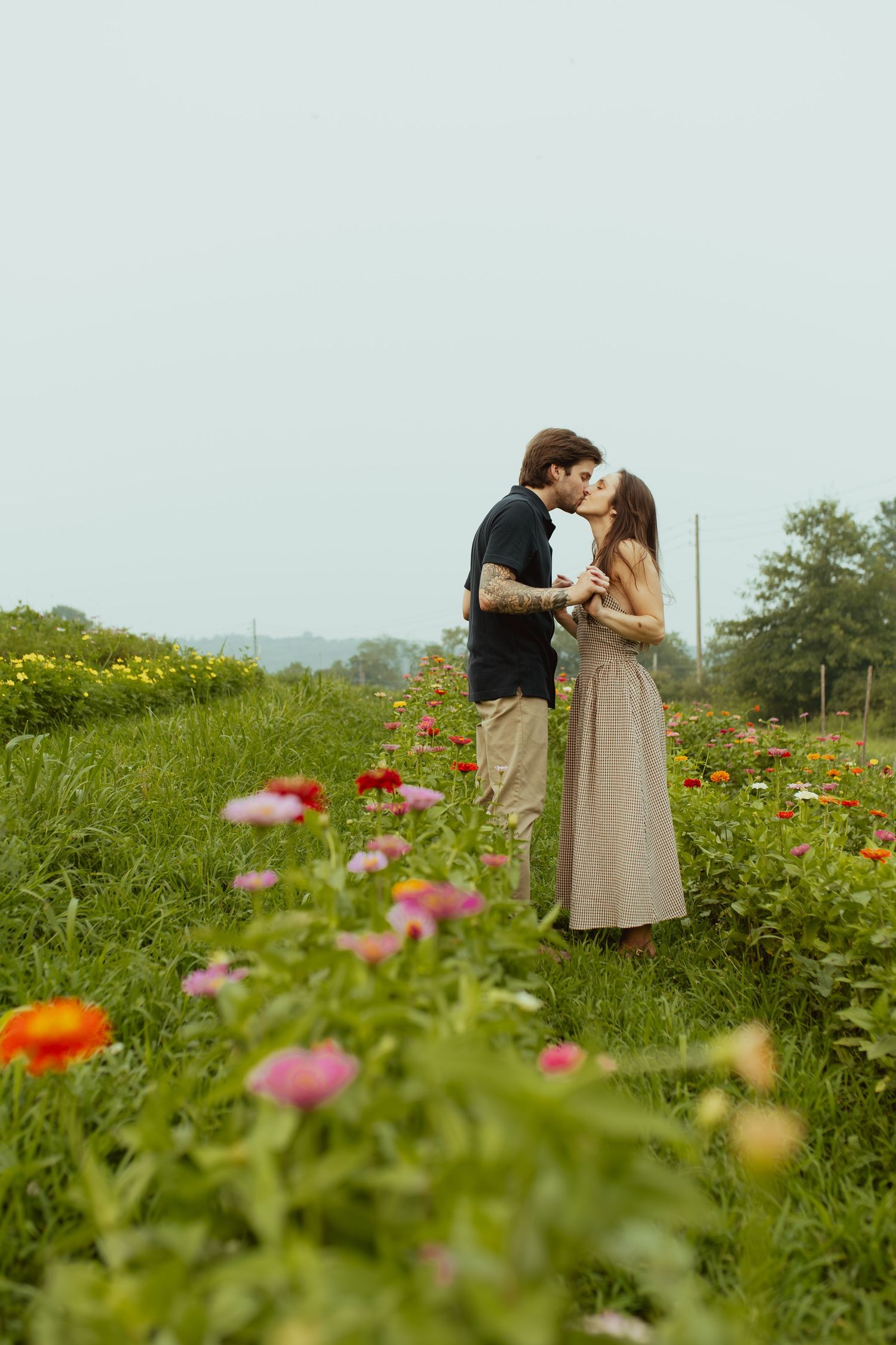 A couple shares a romantic kiss in a vibrant flower field with colorful blooms in the foreground on a cloudy day.