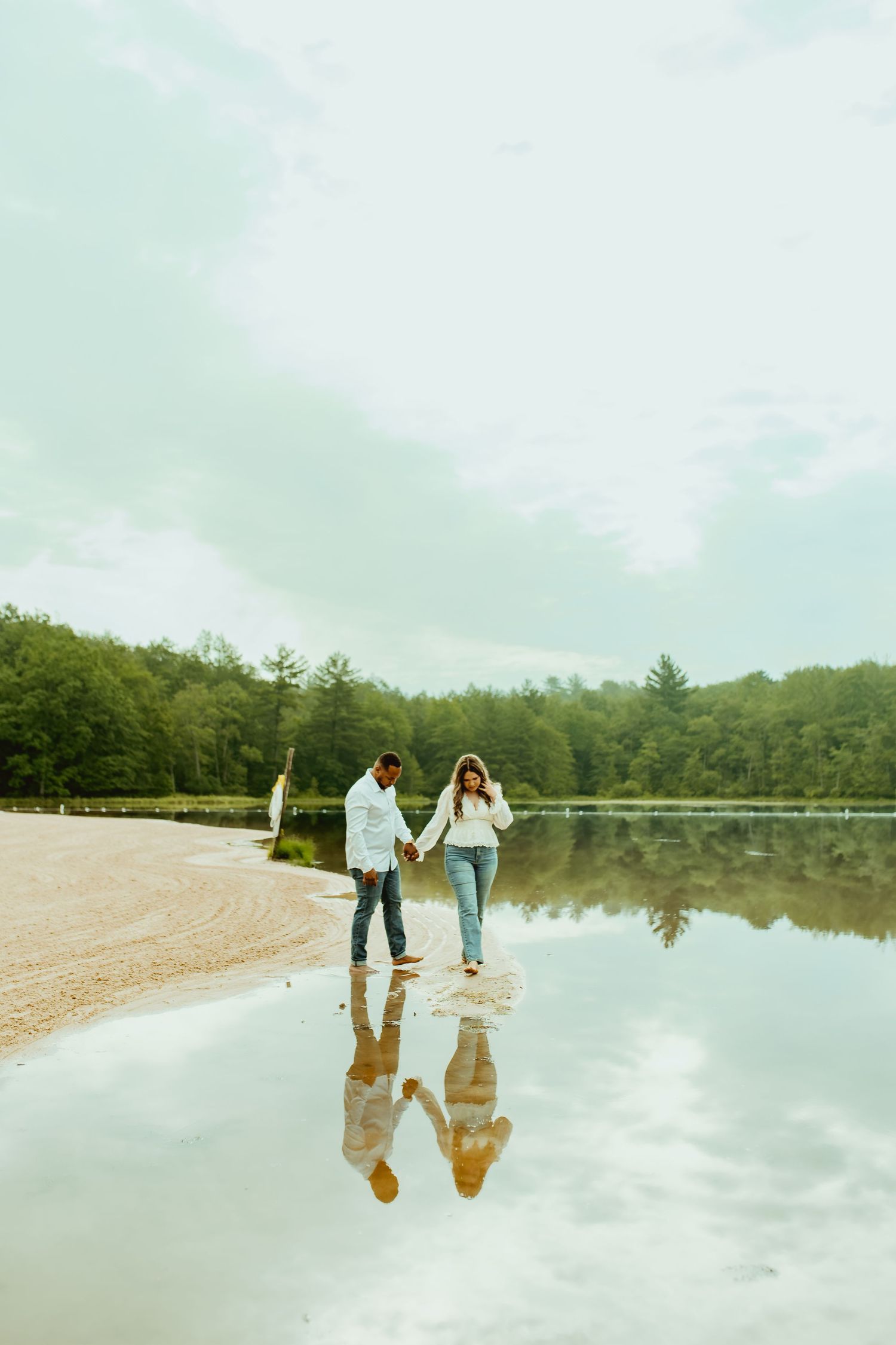 Couple holding hands walking along reflective lake shore with lush green forest backdrop on cloudy day.