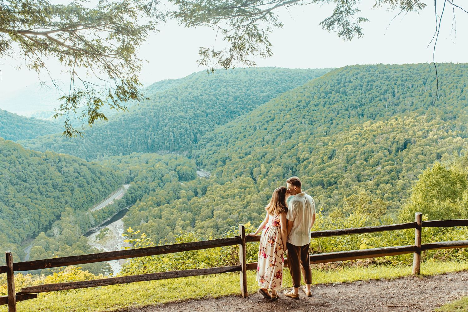 Couple standing at a wooden fence overlook admiring a lush green mountain valley with a winding river below.