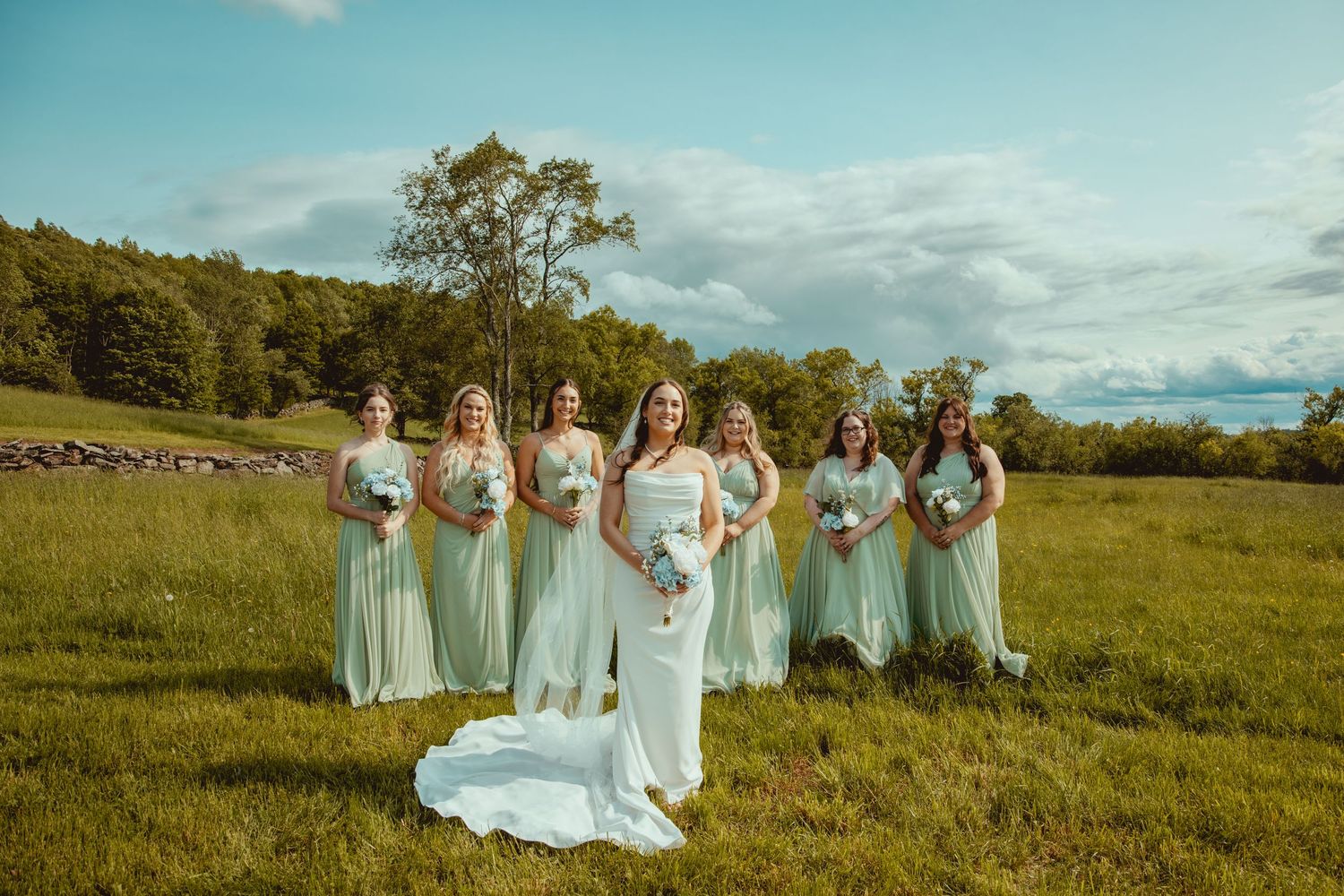 Bridal party in mint green dresses pose with bride in flowing gown against rolling hills.