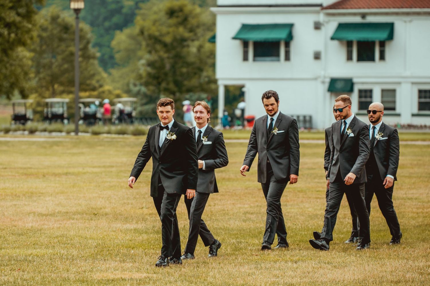 Groomsmen in dark suits walking across a grassy field at an outdoor wedding venue.