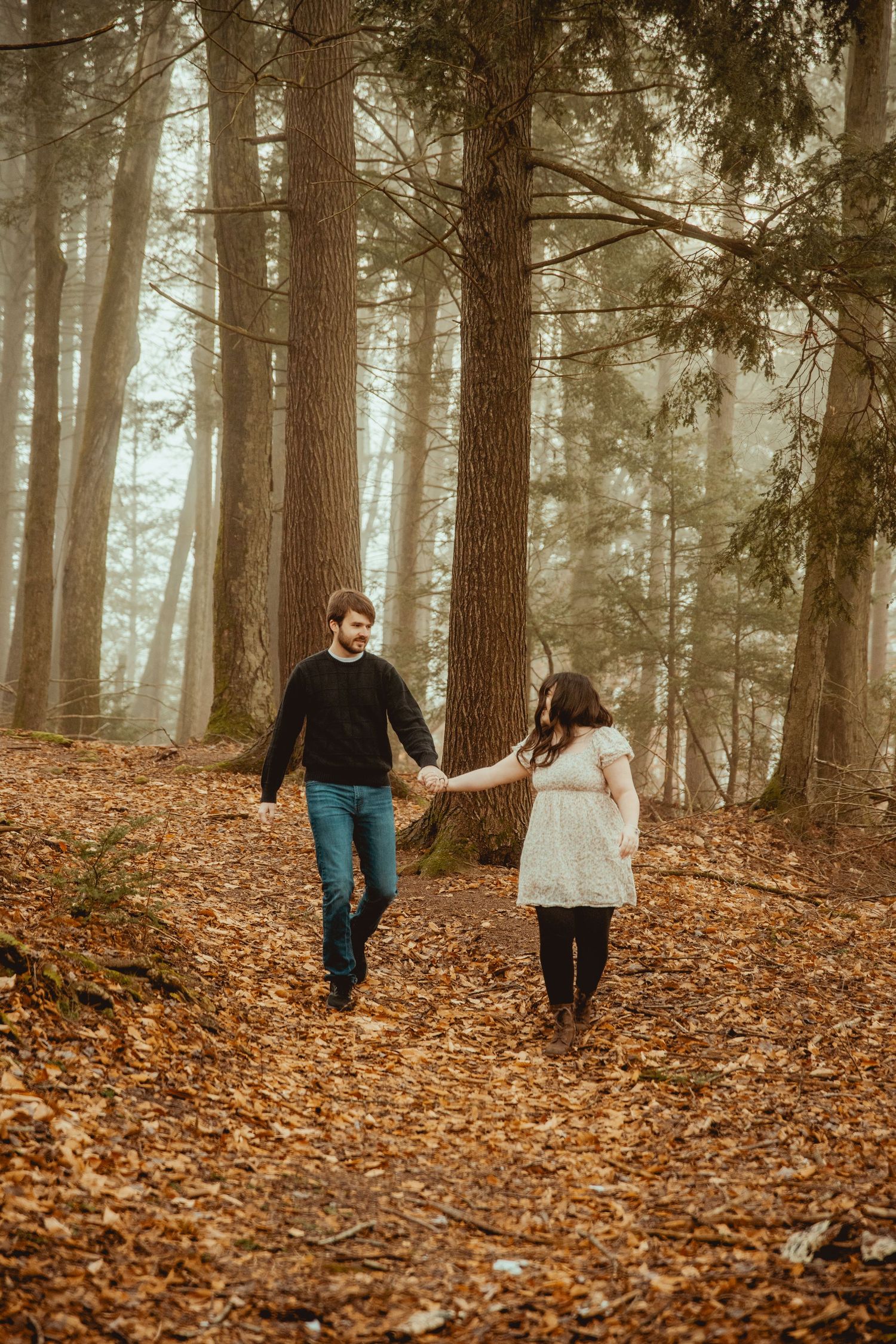 A couple walks hand in hand through a misty autumn forest covered in fallen leaves.