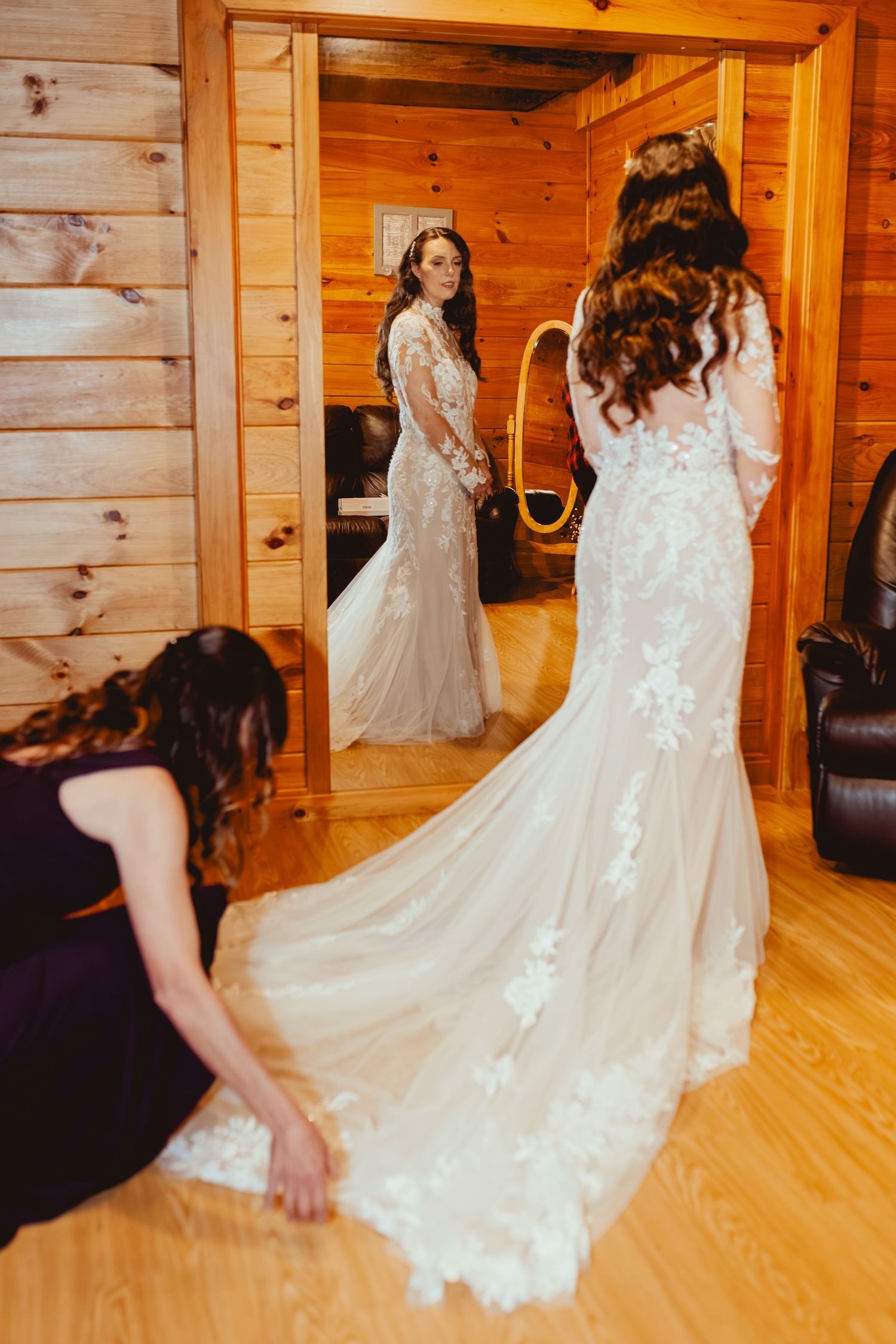 Bride in lace mermaid wedding gown admires reflection in mirror while bridesmaid adjusts train in wooden cabin.