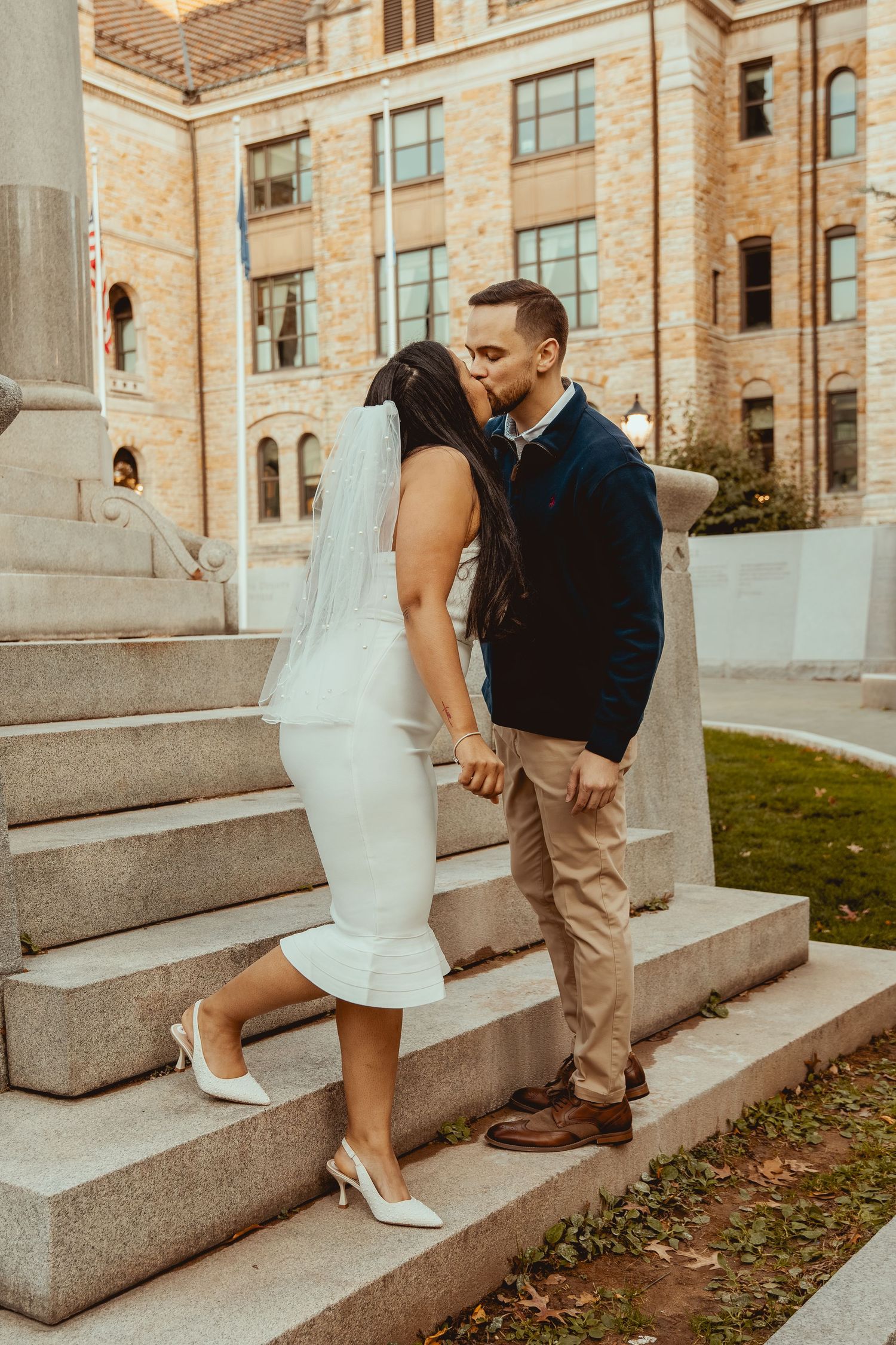 A couple sits on stone steps outside a historic brick building in casual engagement photos.