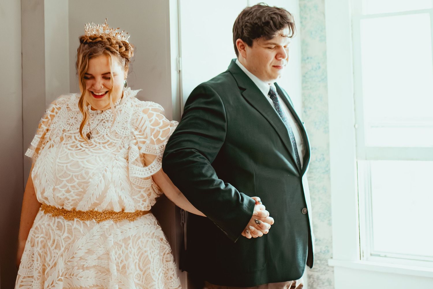 A couple in wedding attire share a tender moment by a bright window in their formal wear.
