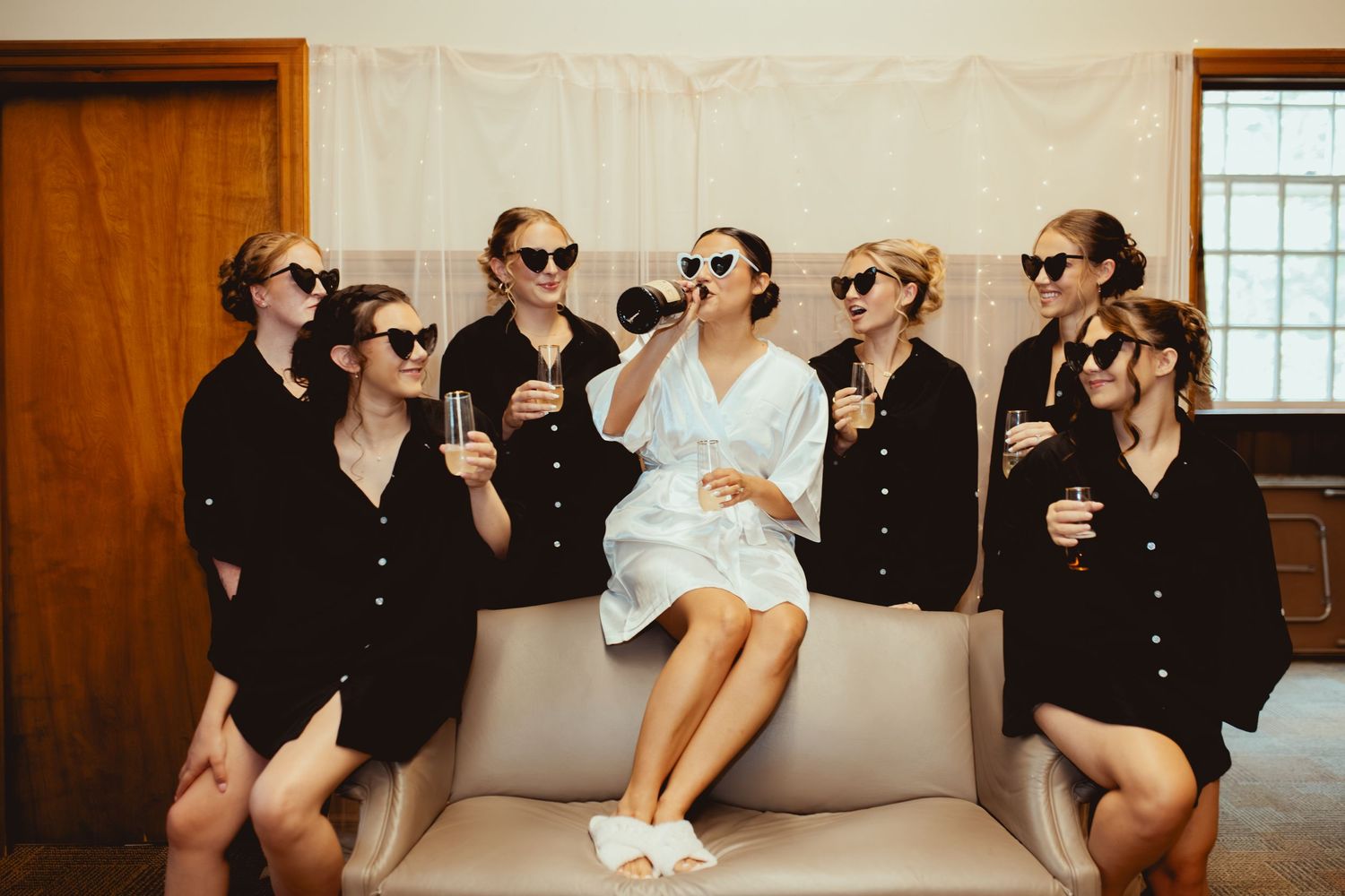 Wedding party members in black attire pose together while drinking from champagne bottles in a rustic indoor setting.