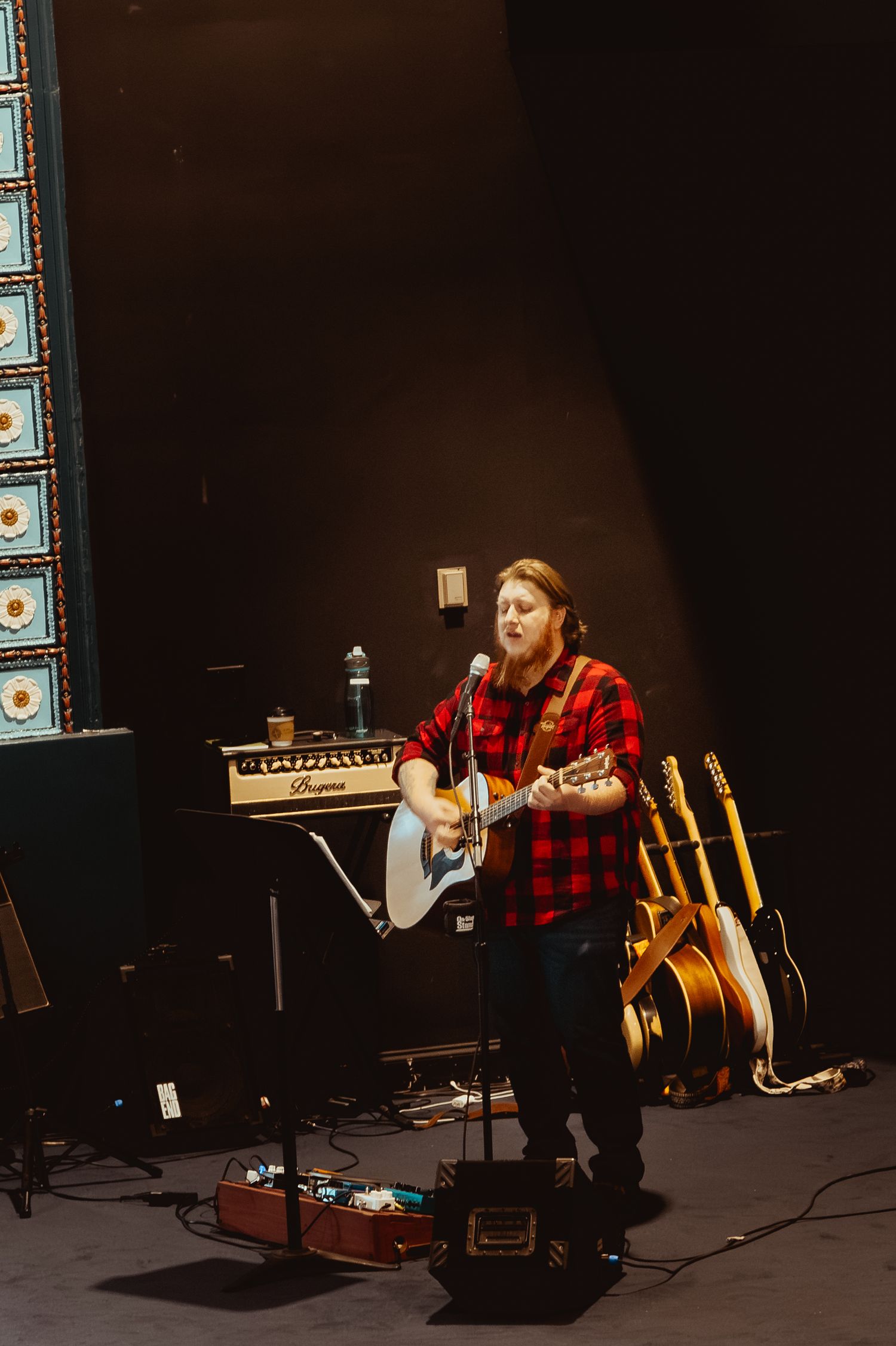 A musician in a red plaid flannel shirt performs on acoustic guitar in a dark performance space.