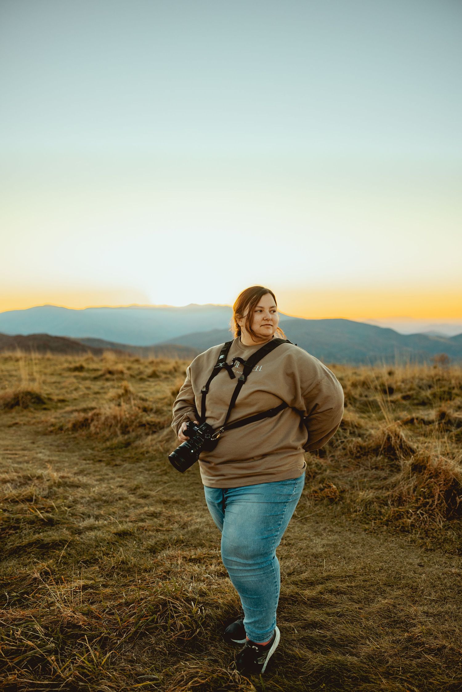 A photographer with a camera stands on a hilltop during golden hour sunset with mountains in the background.