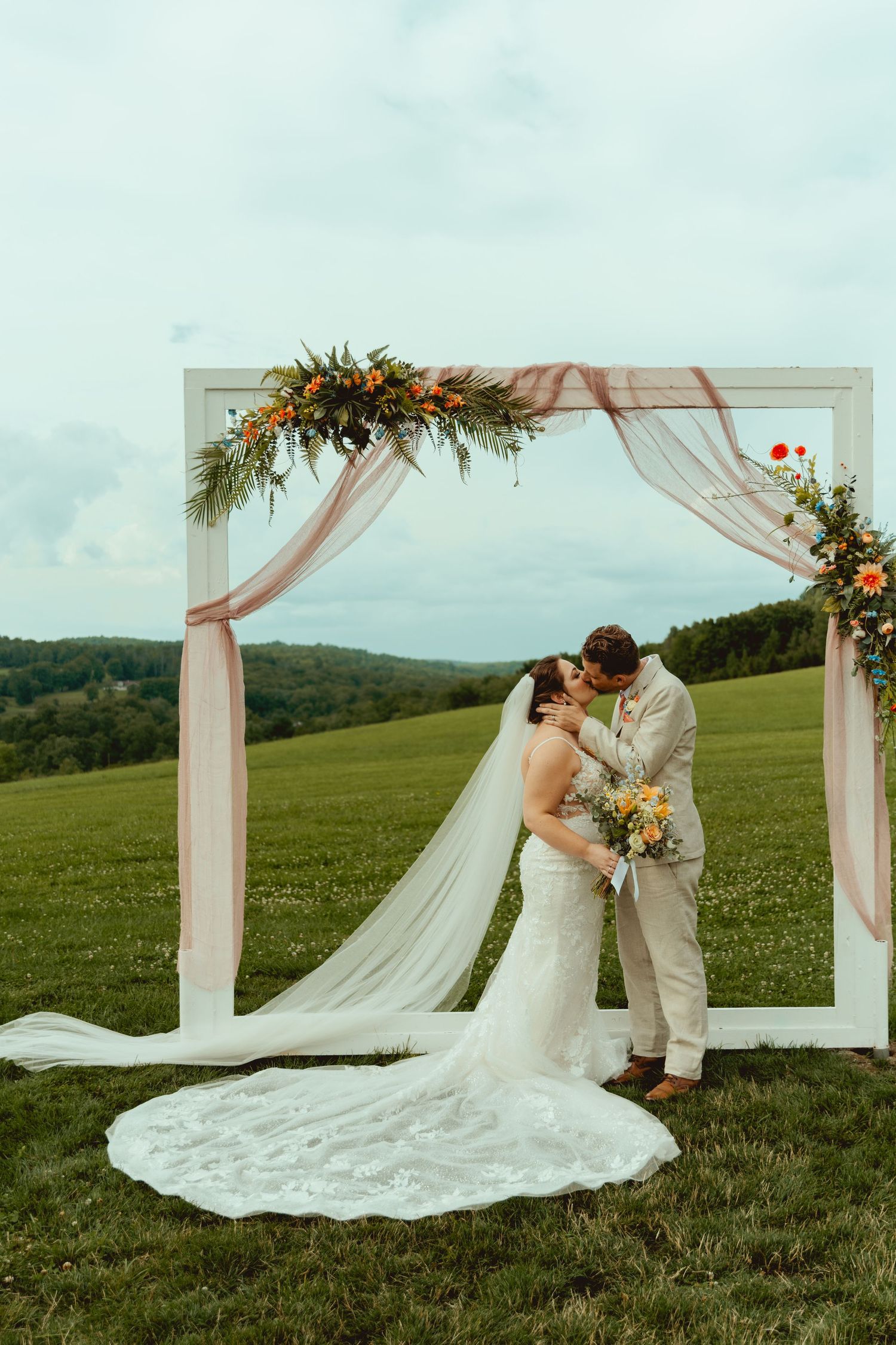 Bride and groom share a kiss under a white floral arch draped with blush fabric on a scenic green meadow.