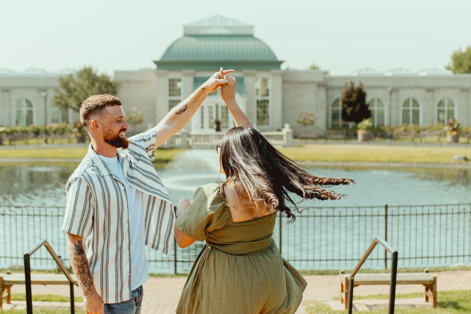 Couple dancing outdoors near a pond with a historic greenhouse building in the background on a sunny day.