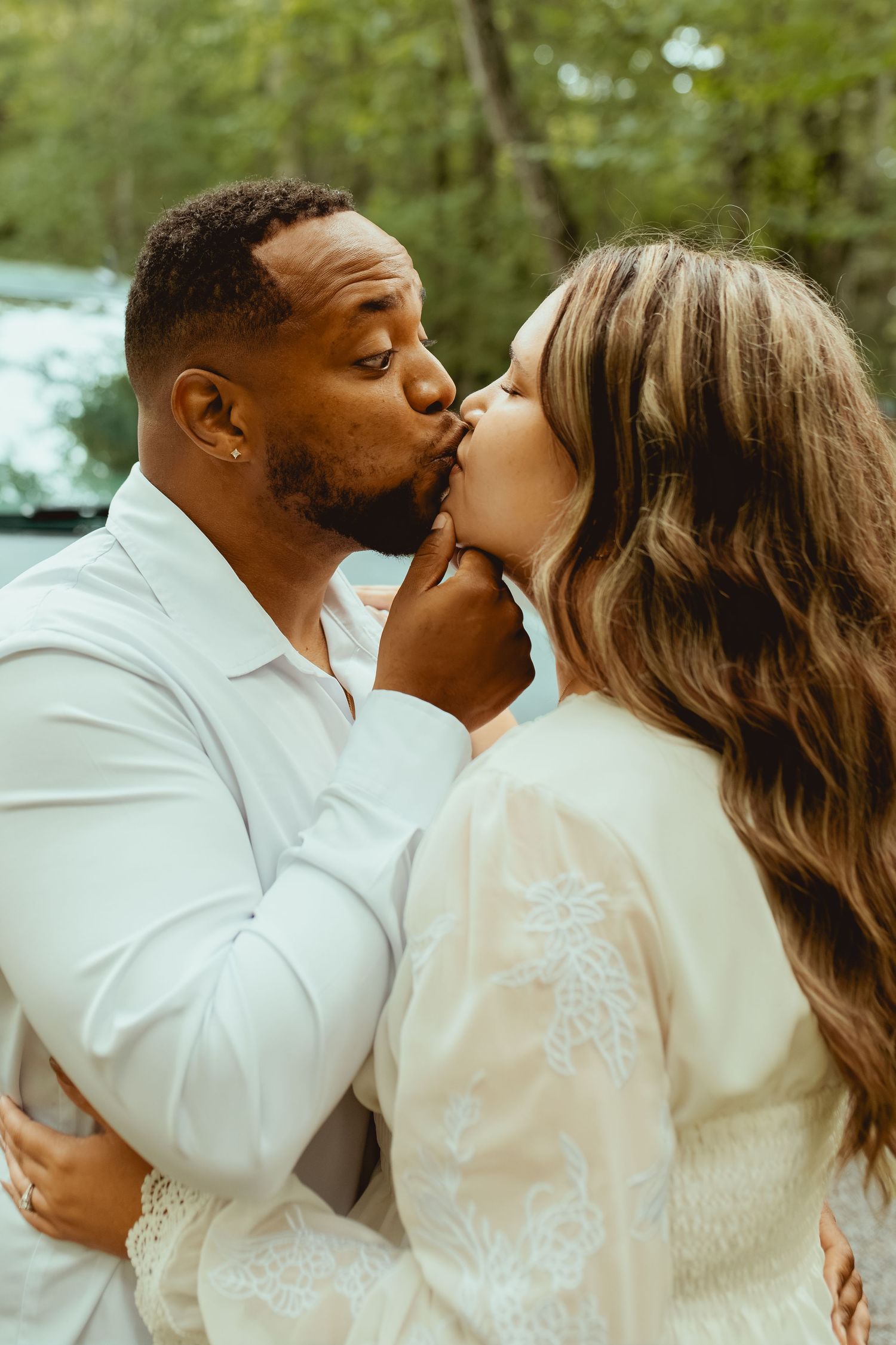 A couple sharing a romantic kiss outdoors, dressed in white, embracing among lush green trees.