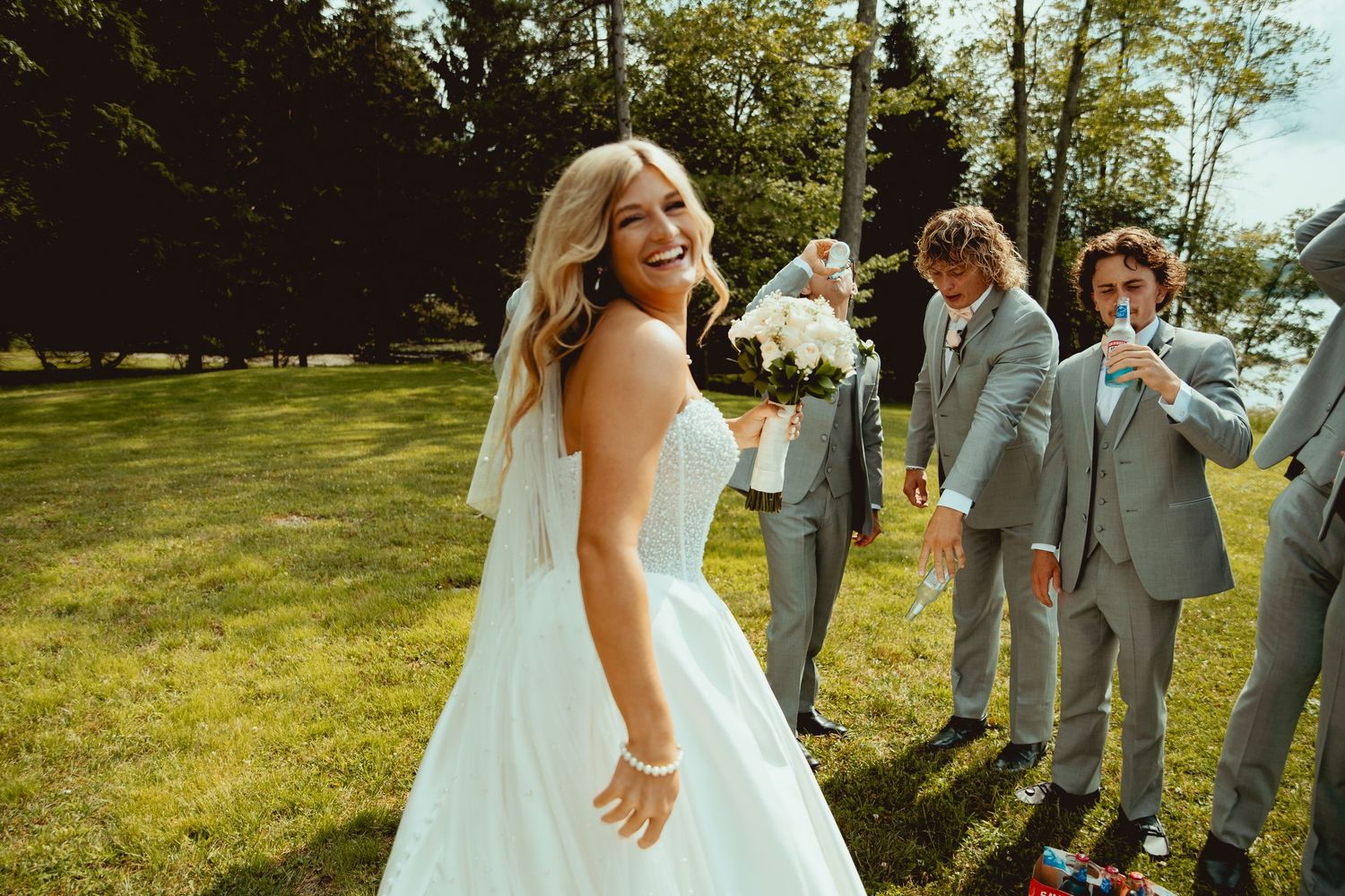Bride in white wedding dress smiling at camera while groomsmen celebrate in background on sunny lawn.