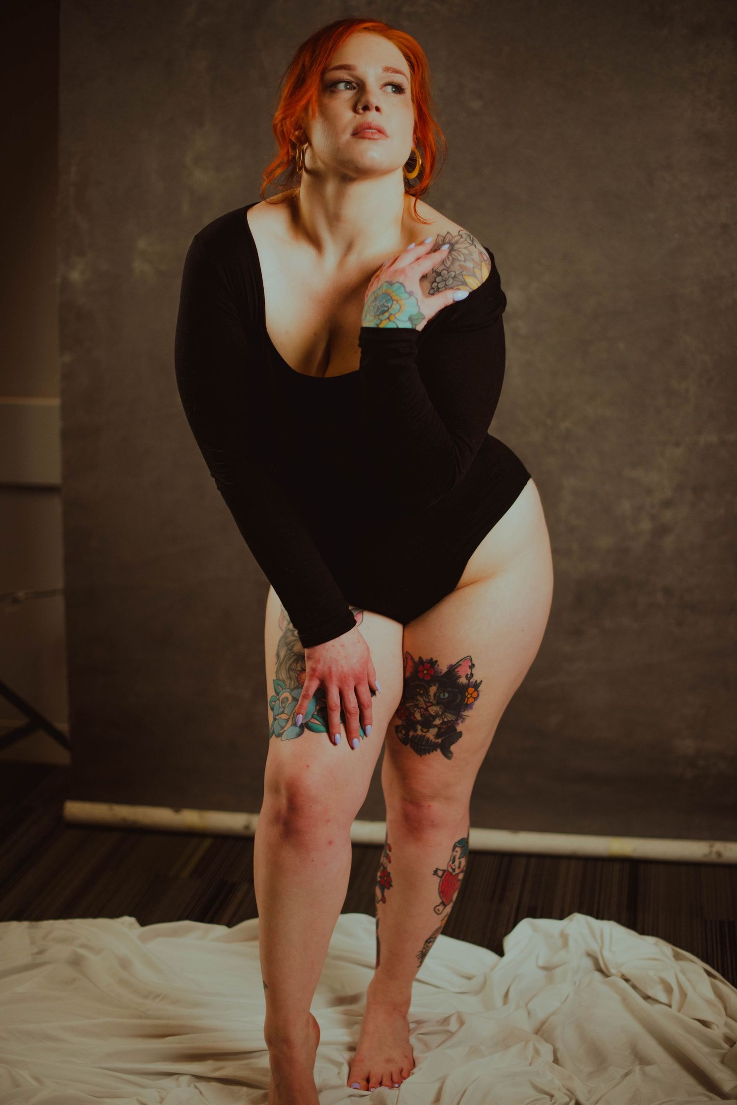 Redhead woman with tattoos poses in black bodysuit against dark studio backdrop.