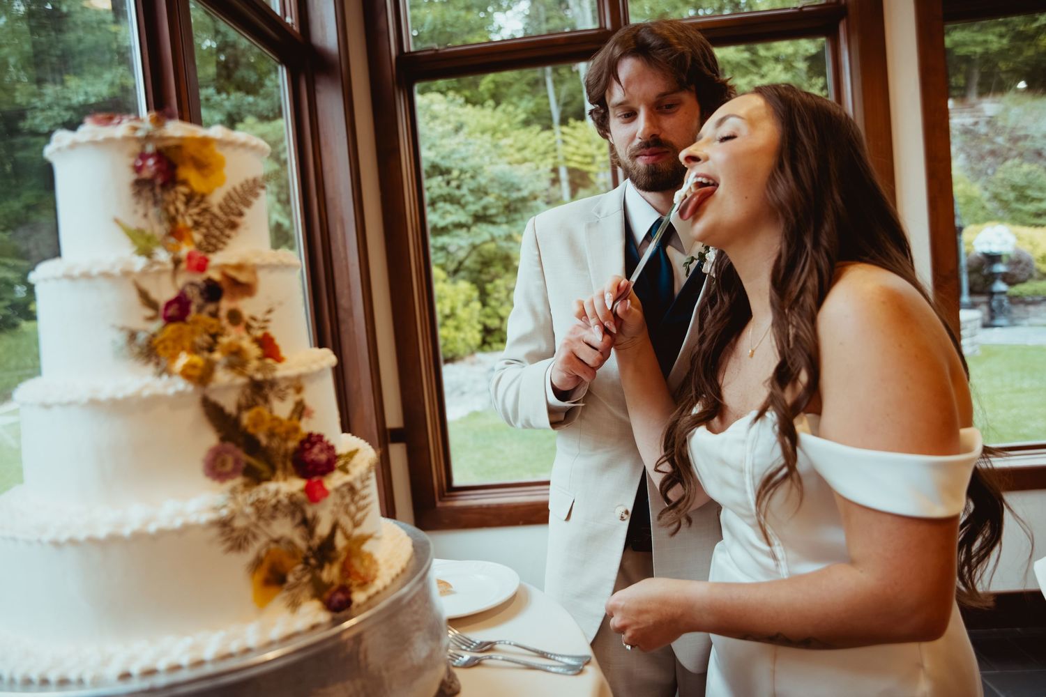 Bride and groom cutting elegant four-tier floral wedding cake indoors at reception venue.