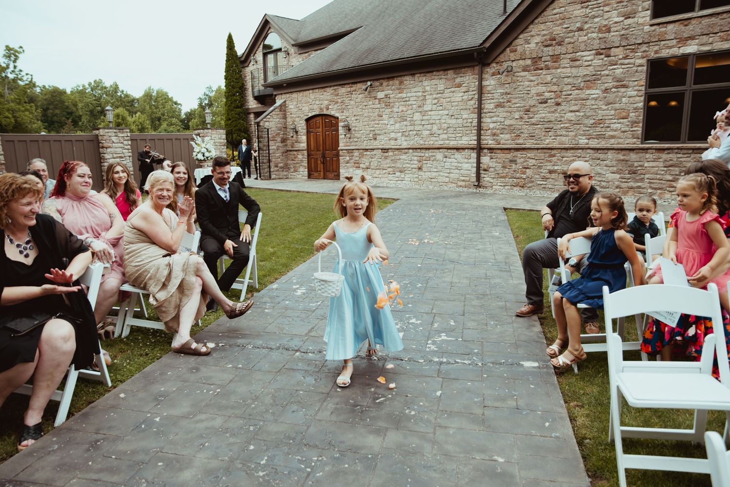 A young flower girl in a blue dress walks down the outdoor wedding aisle as guests watch and smile.