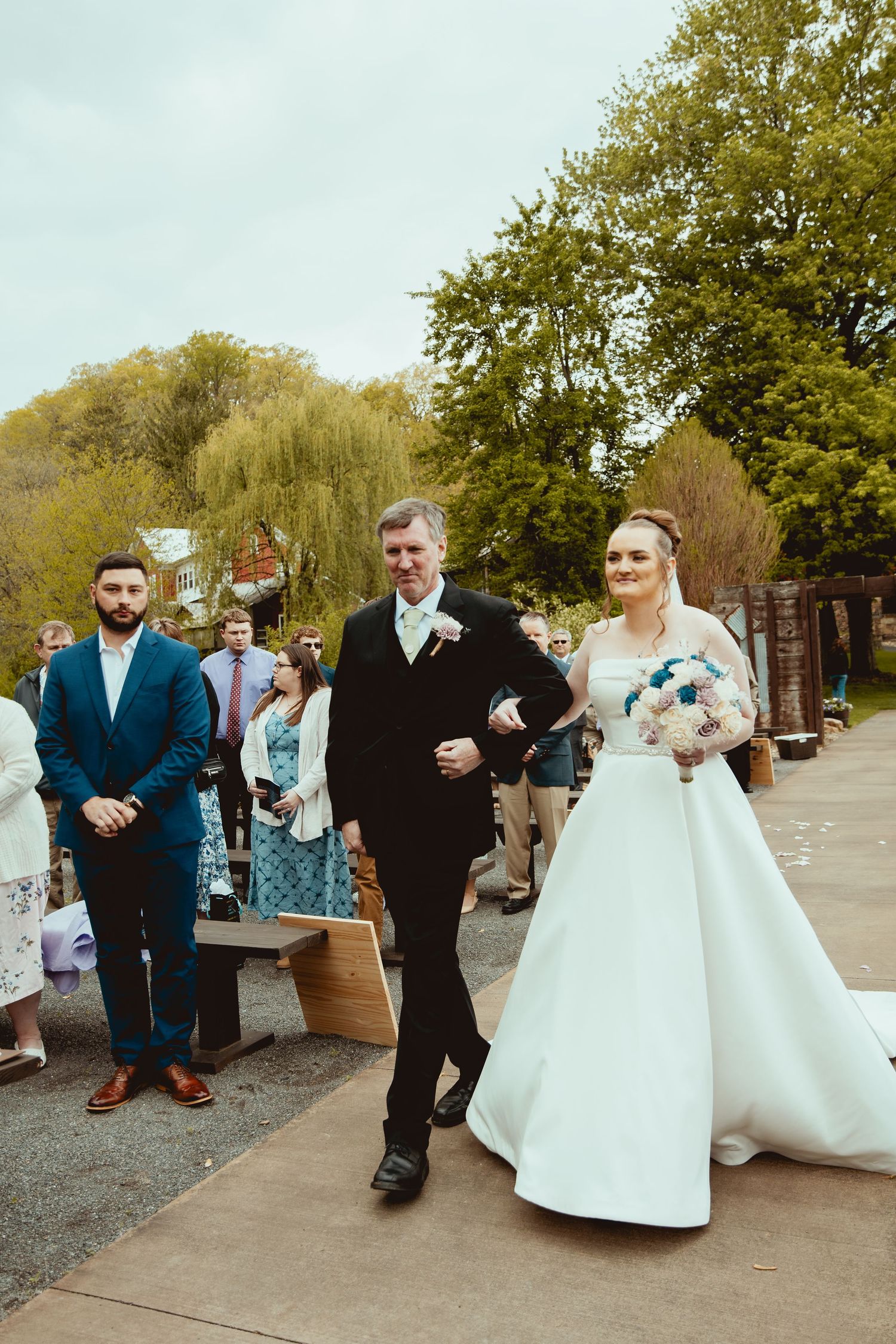 Bride in white ball gown walks down outdoor aisle with escort holding teal bouquet.