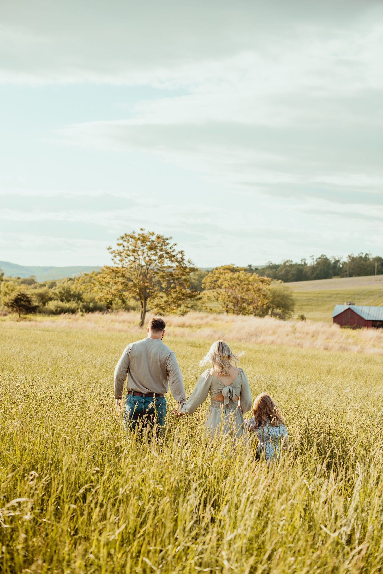 A couple and child walk through a golden wheat field at sunset with mountains in the background.